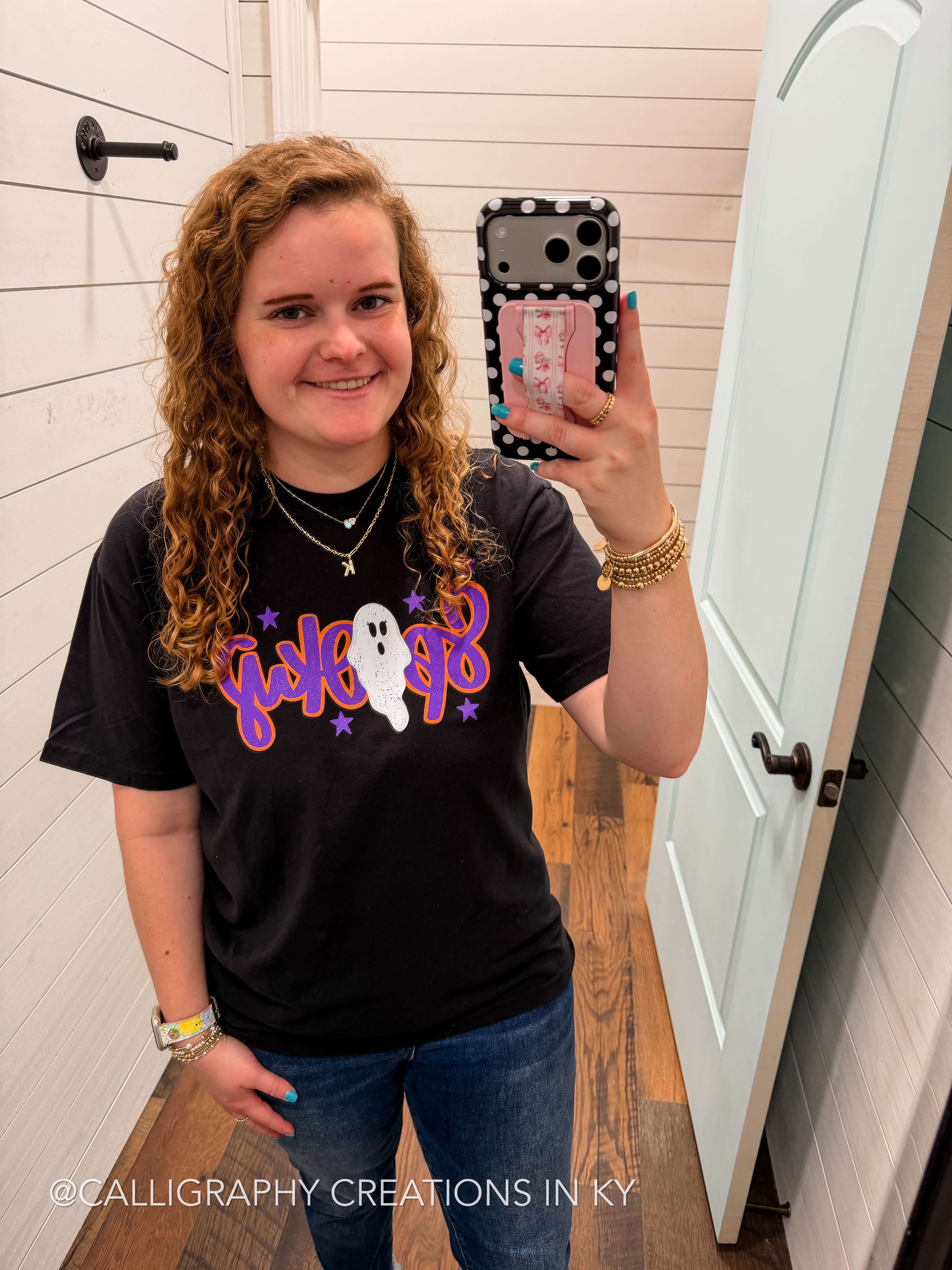 A woman takes a mirror selfie in a bathroom wearing a black graphic t-shirt with a purple and white 'funky ghost' design. The background shows white shiplap walls, a light blue door, and wood flooring.