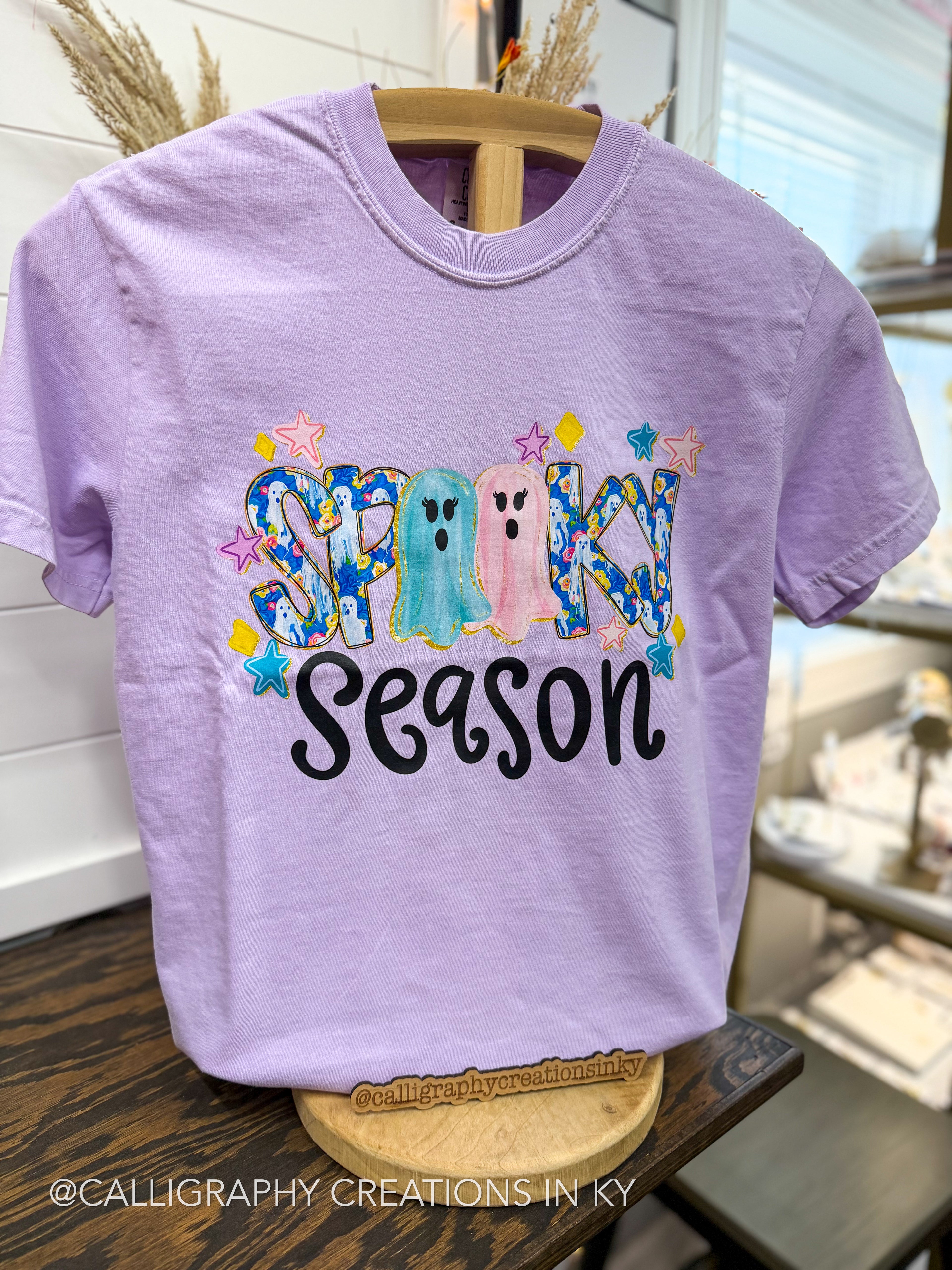 A lavender short-sleeve t-shirt displayed on a wooden hanger in a retail setting. The front features a colorful graphic that reads 'Spooky Season' with two cartoon ghosts and stars.