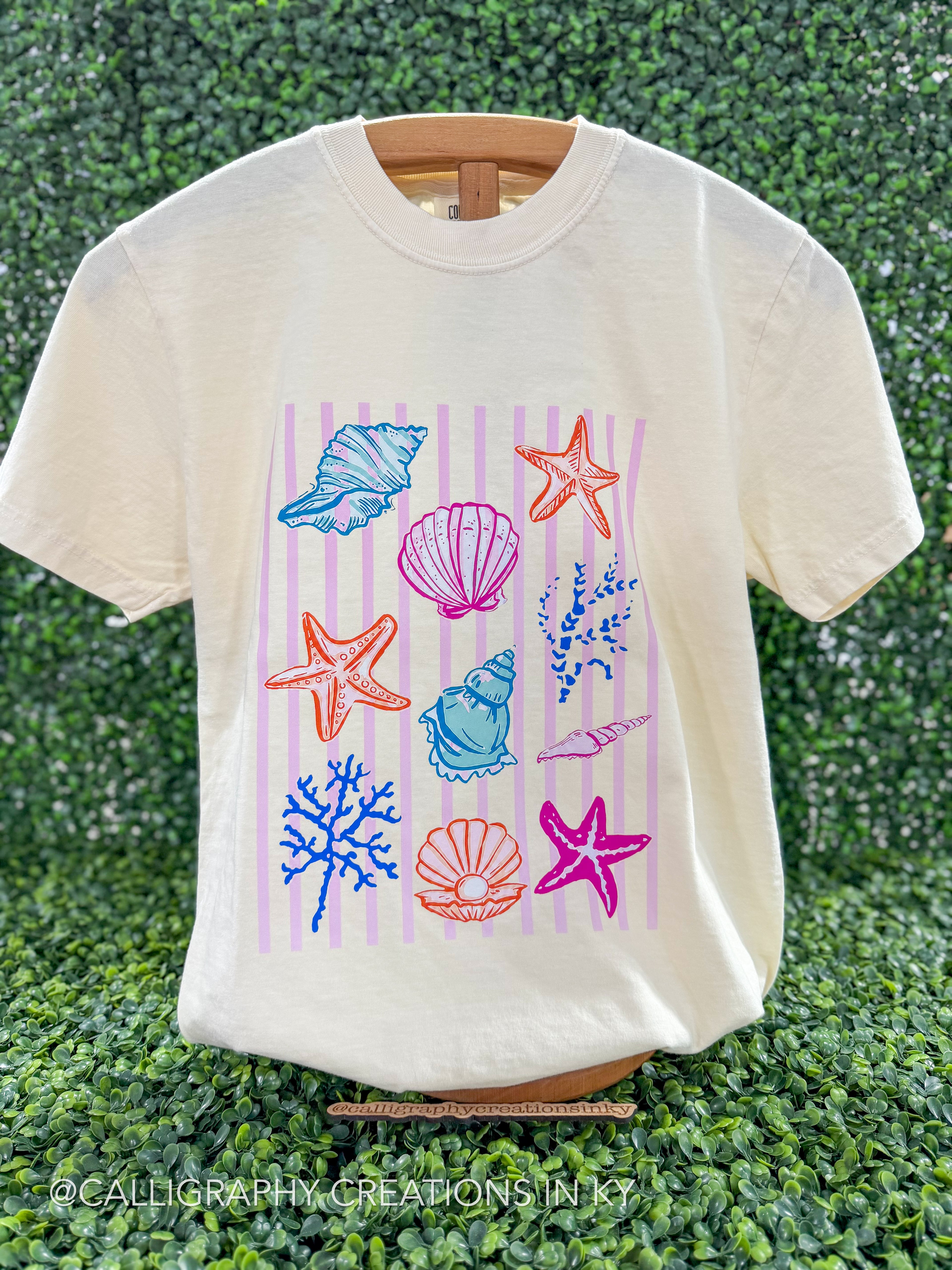 An off-white short-sleeve t-shirt with a colorful seashell and starfish graphic, displayed on a wooden hanger against a green hedge background.