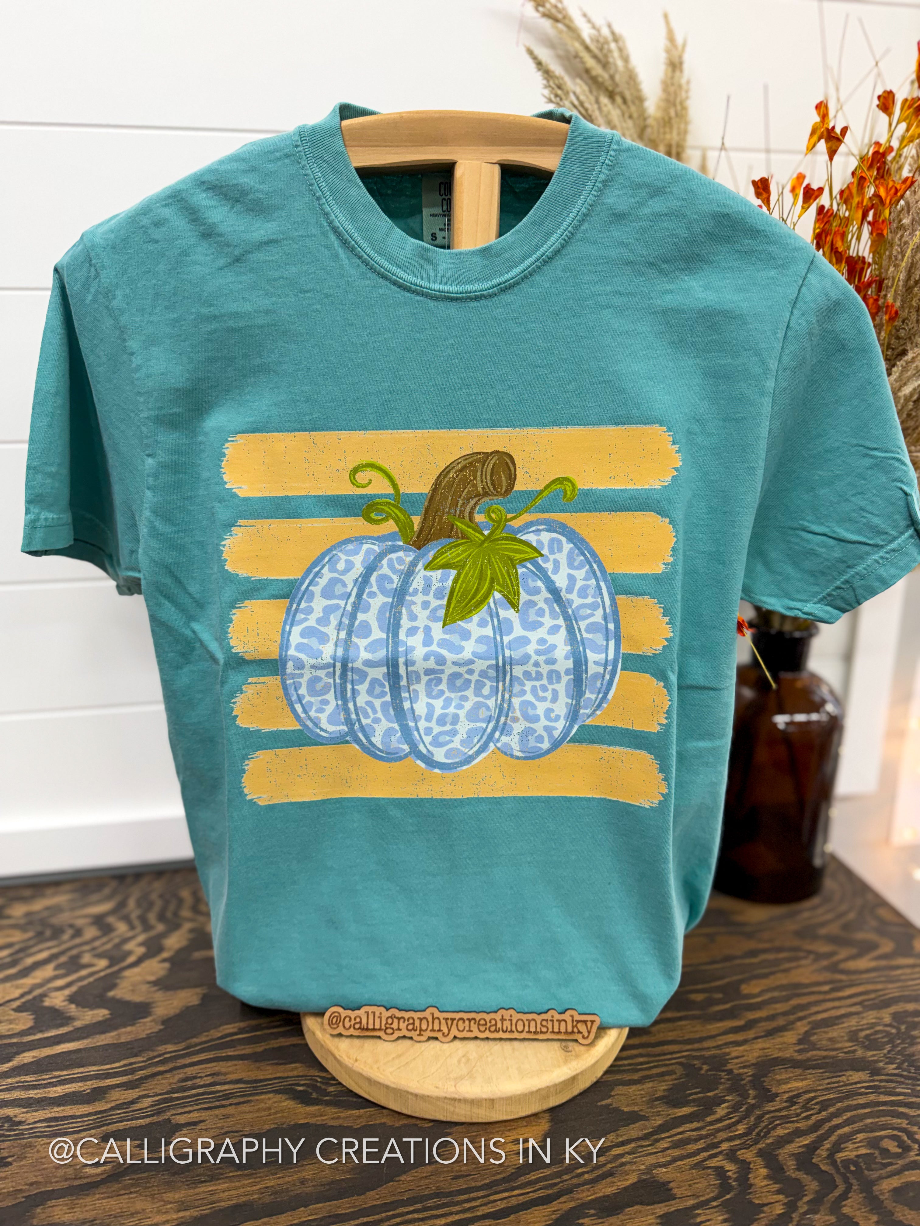 A teal short-sleeve t-shirt displayed on a wooden hanger stand, featuring a graphic of a blue patterned pumpkin with a green stem and brown stem, set against yellow horizontal stripes.
