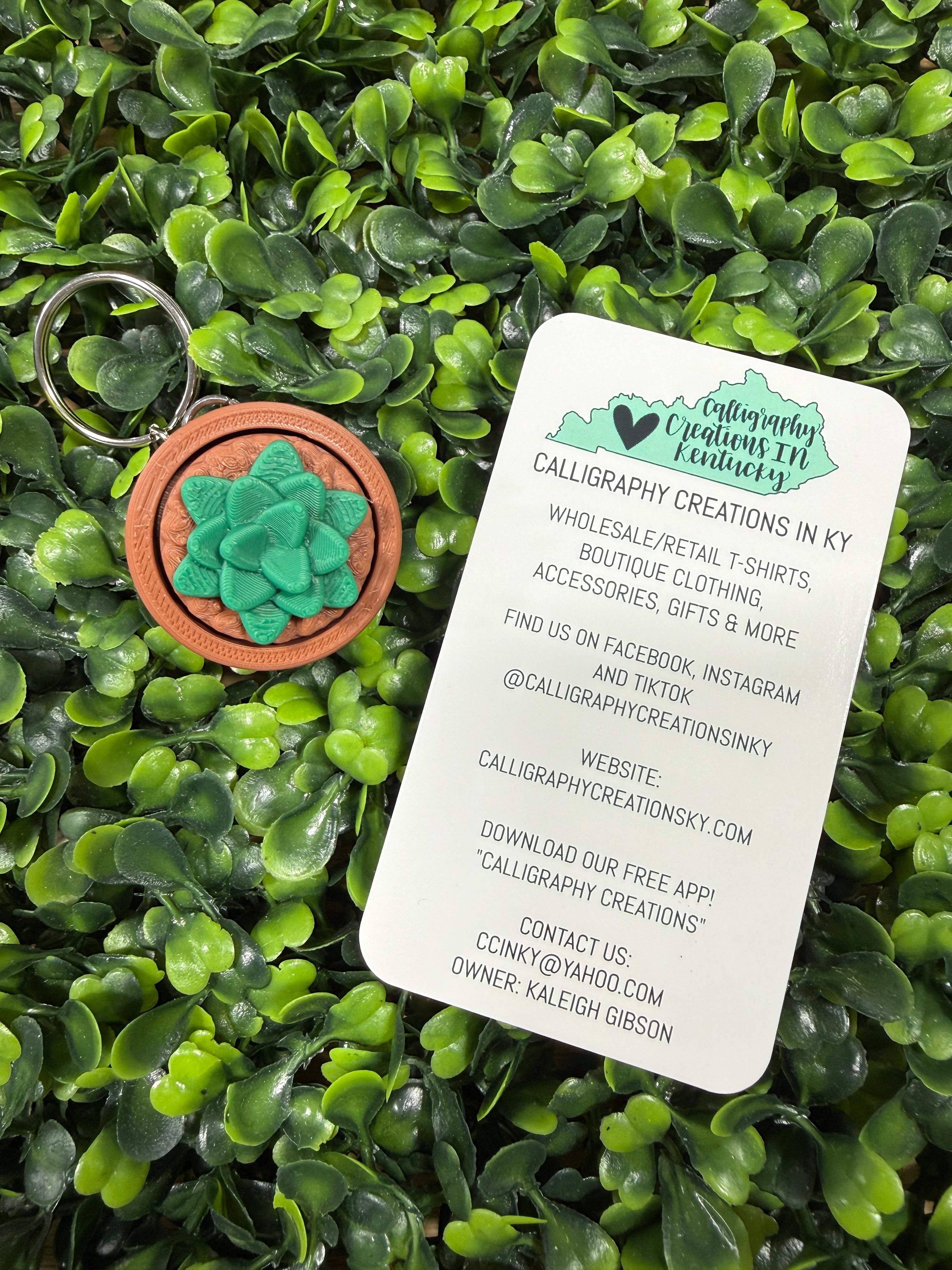 A circular wooden keychain with a green succulent design and a split key ring, placed on a bed of green artificial leaves next to a white business card for 'Caligraphy Creations in KY'.