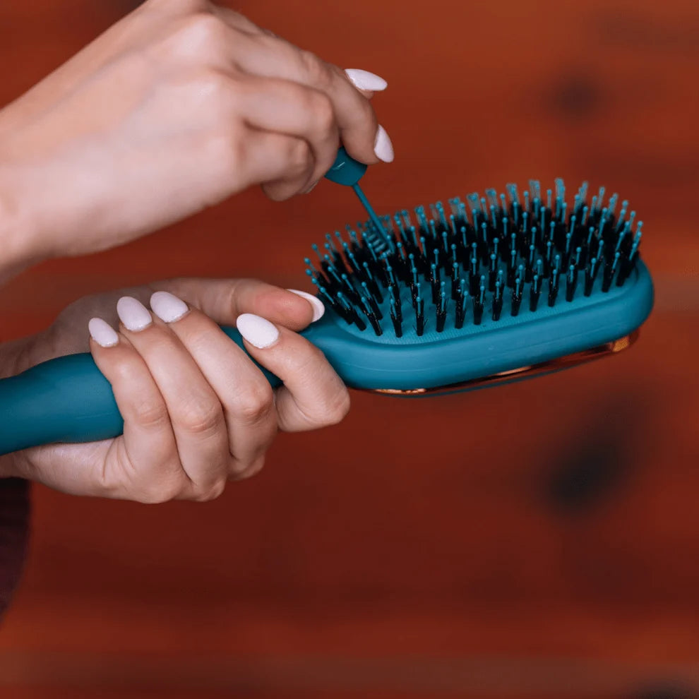 A close-up of a person's hands holding a teal hairbrush while applying a small blue hair product from a tube. The background is a warm, blurred reddish-brown surface.