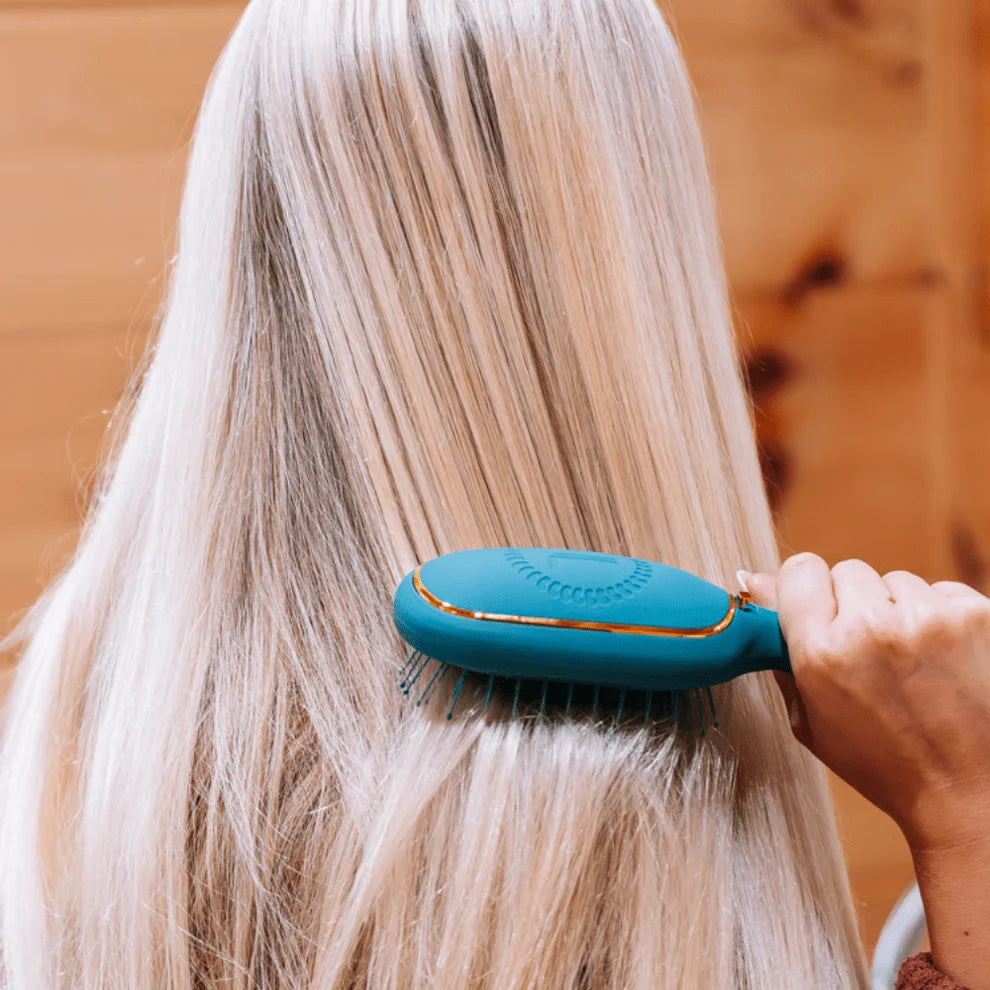 A close-up lifestyle shot of a person with long, straight platinum blonde hair using a teal and gold hairbrush to brush their hair. The background is a soft-focus warm wood panel.