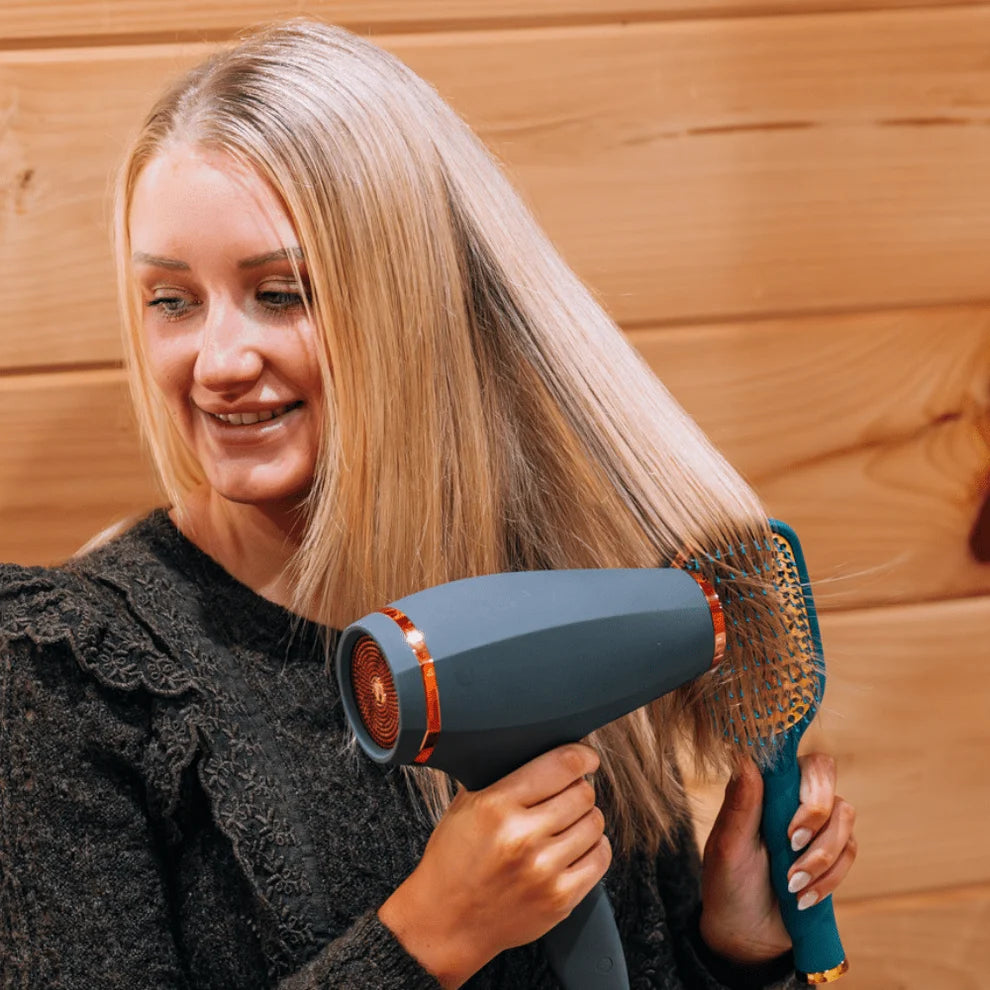 A woman with long blonde hair uses a gray hair dryer with a teal and gold paddle brush attachment against her hair, set against a warm wood-paneled background.
