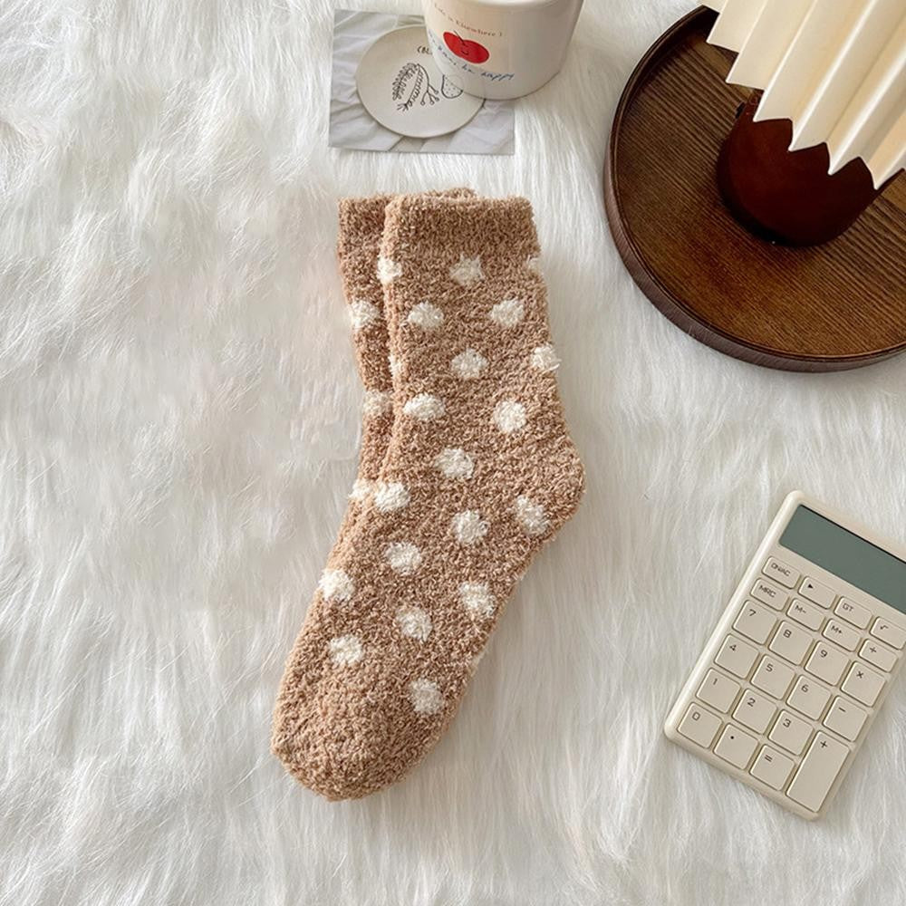 A pair of plush ankle socks in a warm taupe-brown color with white polka dots, laid flat on a white faux fur surface with styled props including a wooden tray, a calculator, and a mug.