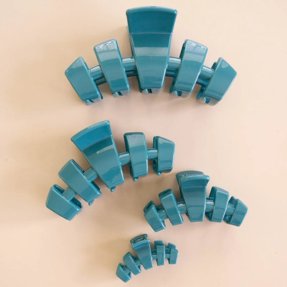Four glossy teal hair claw clips in different sizes arranged on a light beige background.