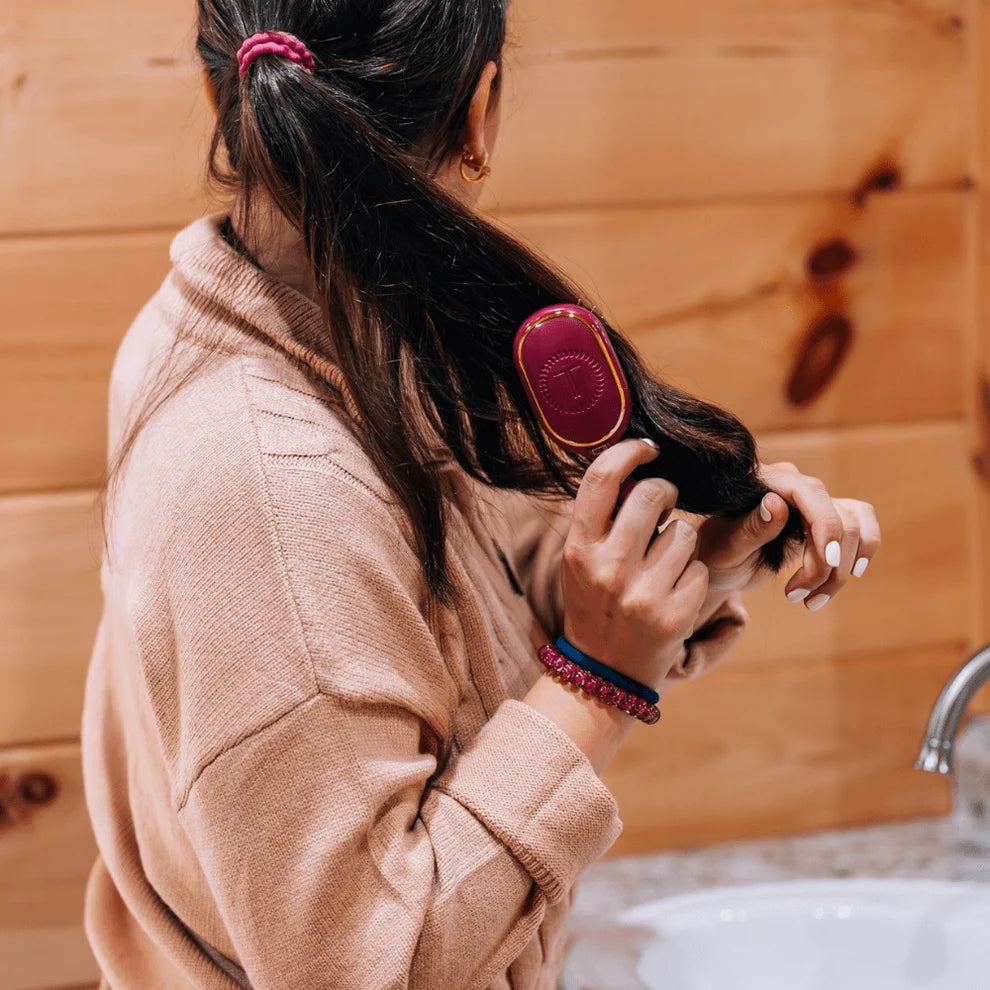 A woman with dark hair tied back in a ponytail uses a pink and gold hairbrush on her hair, standing in front of a wooden wall with a sink visible.