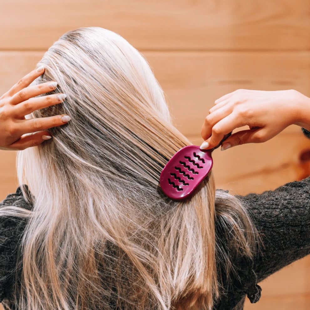 A person with long blonde hair seen from behind, using a dark pink hairbrush to brush their hair, with a warm wood-paneled background.