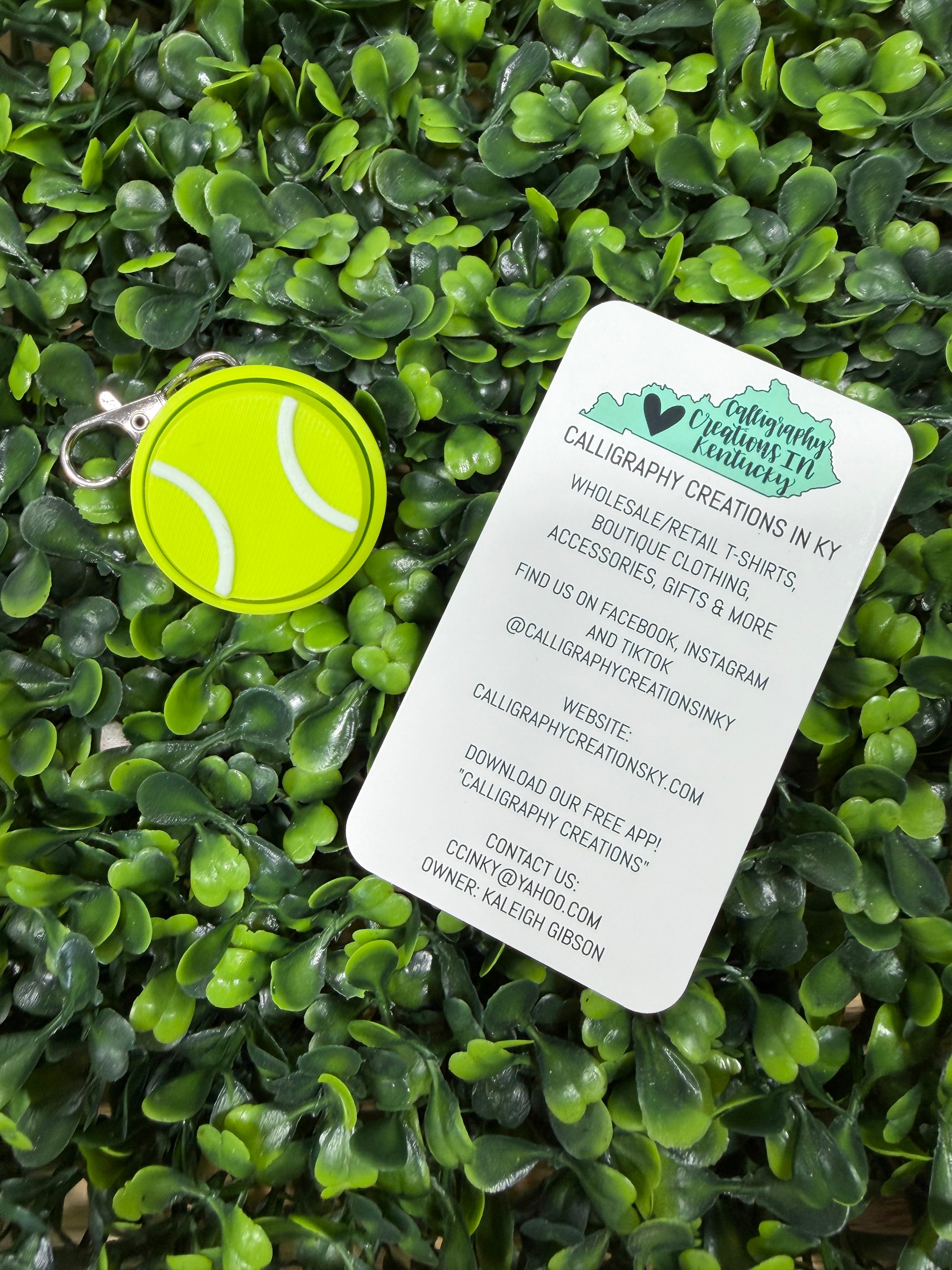 A bright yellow-green tennis ball-shaped keychain with a silver metal ring lies on a white printed business card, both placed on dense green foliage. The card shows the brand Calligraphy Creations in Ky and contact details.