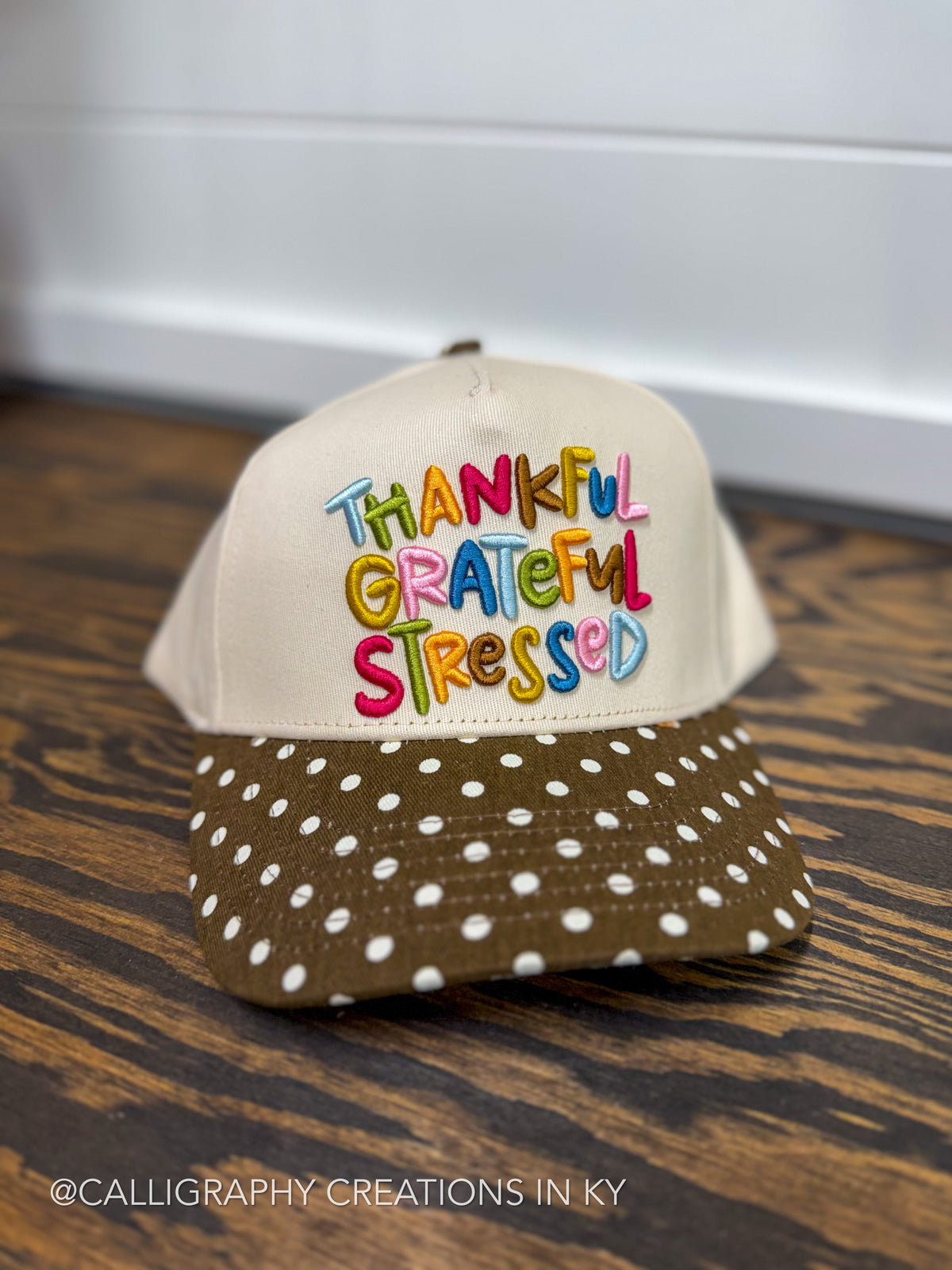 A beige trucker-style cap with colorful embroidered text reading 'THANKFUL GRATEFUL STRESSED' on the front, a brown brim with white polka dots, and a wood-grain patterned underside, displayed on a dark wooden surface.