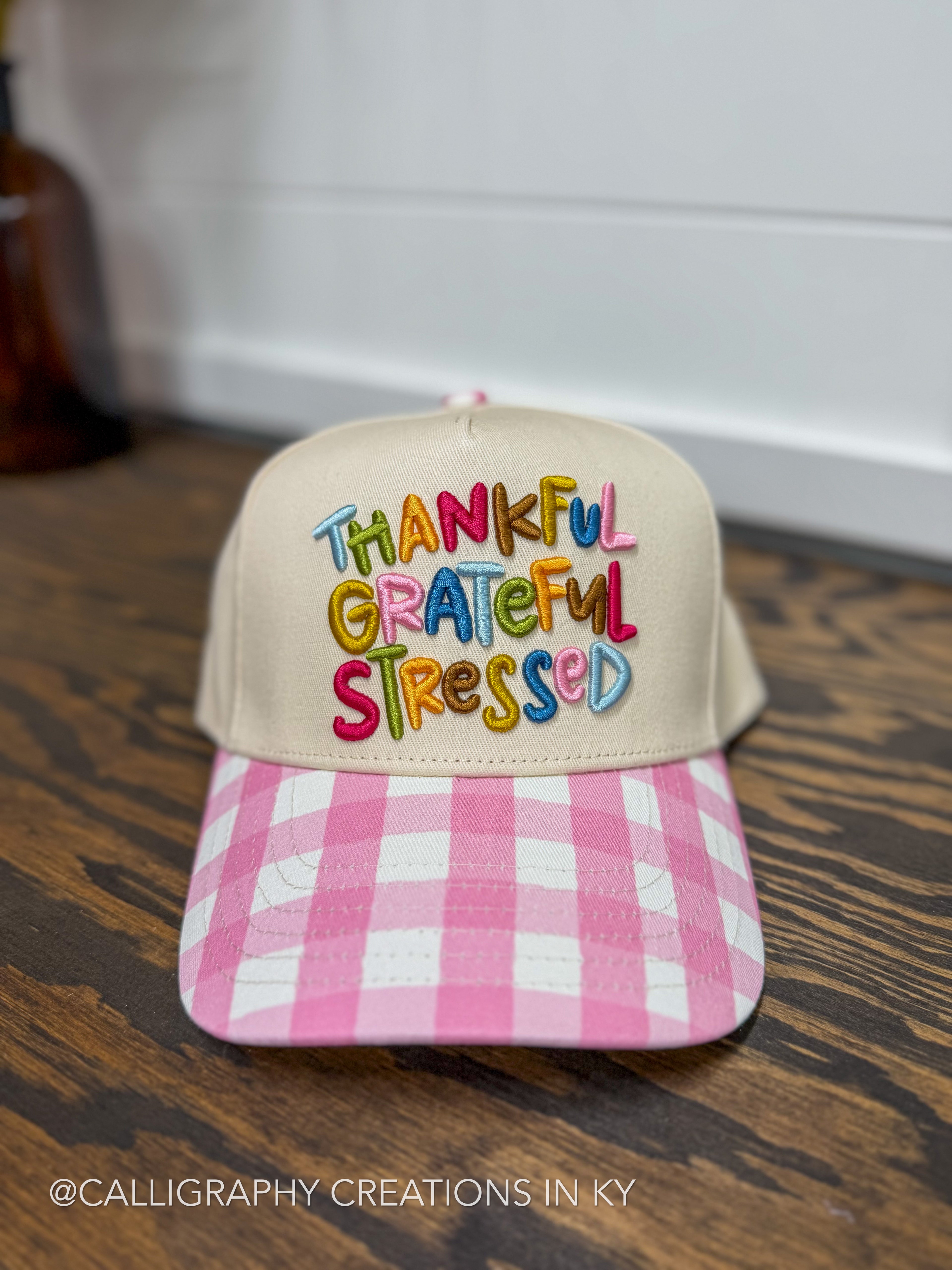 A beige baseball cap with colorful embroidered text reading 'THANKFUL GRATEFUL STRESSED' on the front, a pink and white gingham patterned brim, and a wooden tabletop background.