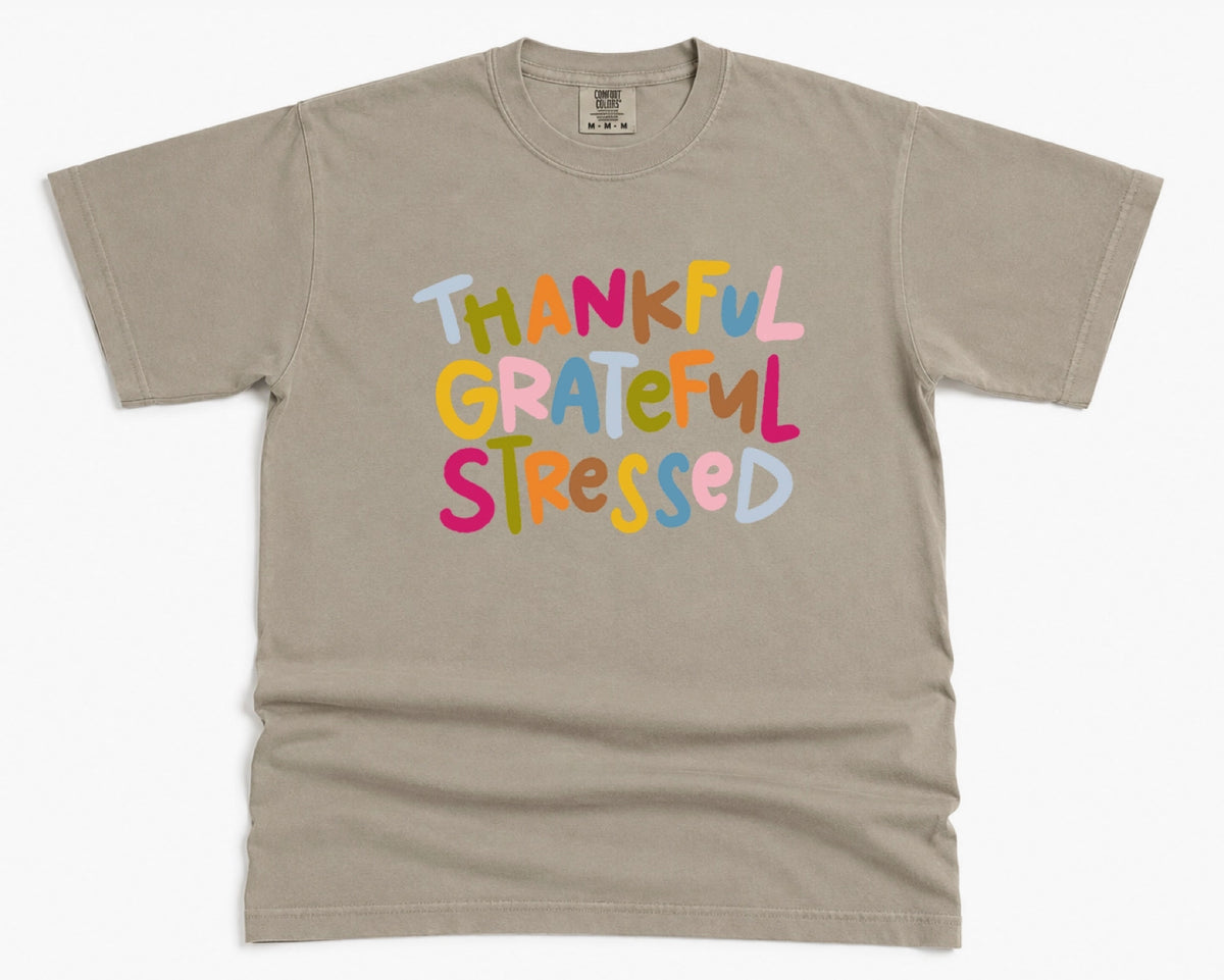 A taupe gray short-sleeve t-shirt with the phrase 'THANKFUL GRATEFUL STRESSED' printed on the chest in colorful, playful bubble letters.