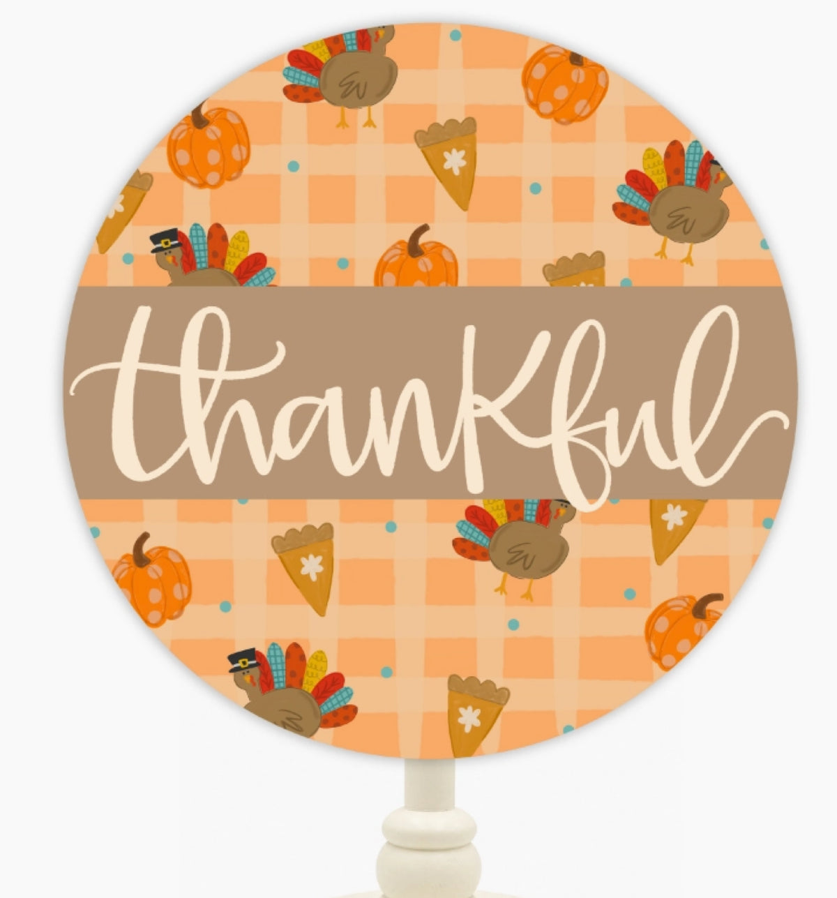 A round sign with a Thanksgiving theme, featuring a pattern of pumpkins, turkeys, and corn on a peach and orange plaid background, with a brown banner across the middle that reads 'thankful' in white script. The sign is mounted on a white post.