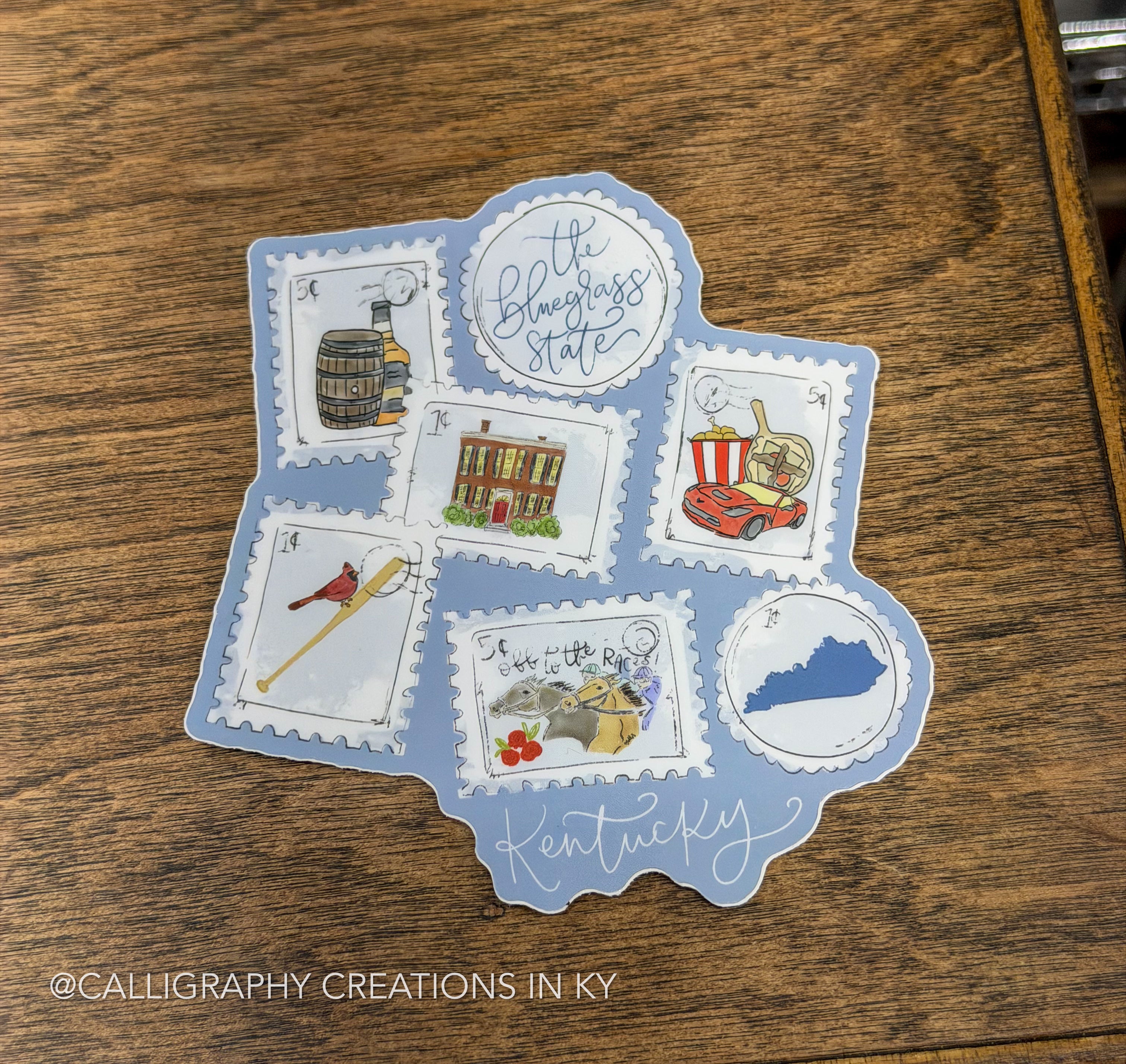 A Kentucky-themed calligraphy sticker or decal with a light blue background and six postage-stamp-style illustrations, placed on a wooden tabletop. The design includes a barrel, a church, a popcorn bucket, a rifle, flowers, a blue silhouette of Kentucky, and the words 'The Bluegrass State' and 'Kentucky.