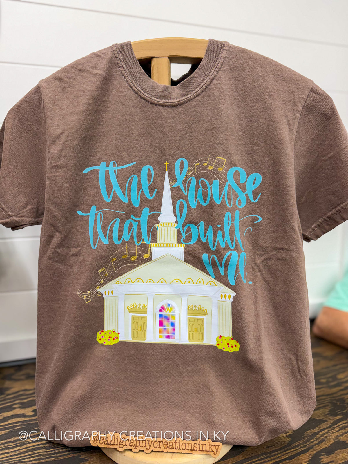 A brown short-sleeve t-shirt on a wooden hanger, featuring a graphic of a white church with a tall steeple and the phrase 'the house that built me' in light blue cursive script.
