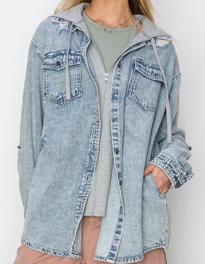 A woman wearing an oversized light-wash blue denim hooded jacket with distressed details, layered over a sage green top and light pink pants, against a plain white background.