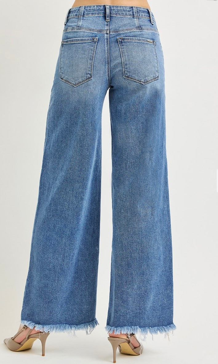 Wide-leg blue denim jeans with a frayed hem, shown from the back on a model wearing beige high-heeled sandals against a light gray studio background.