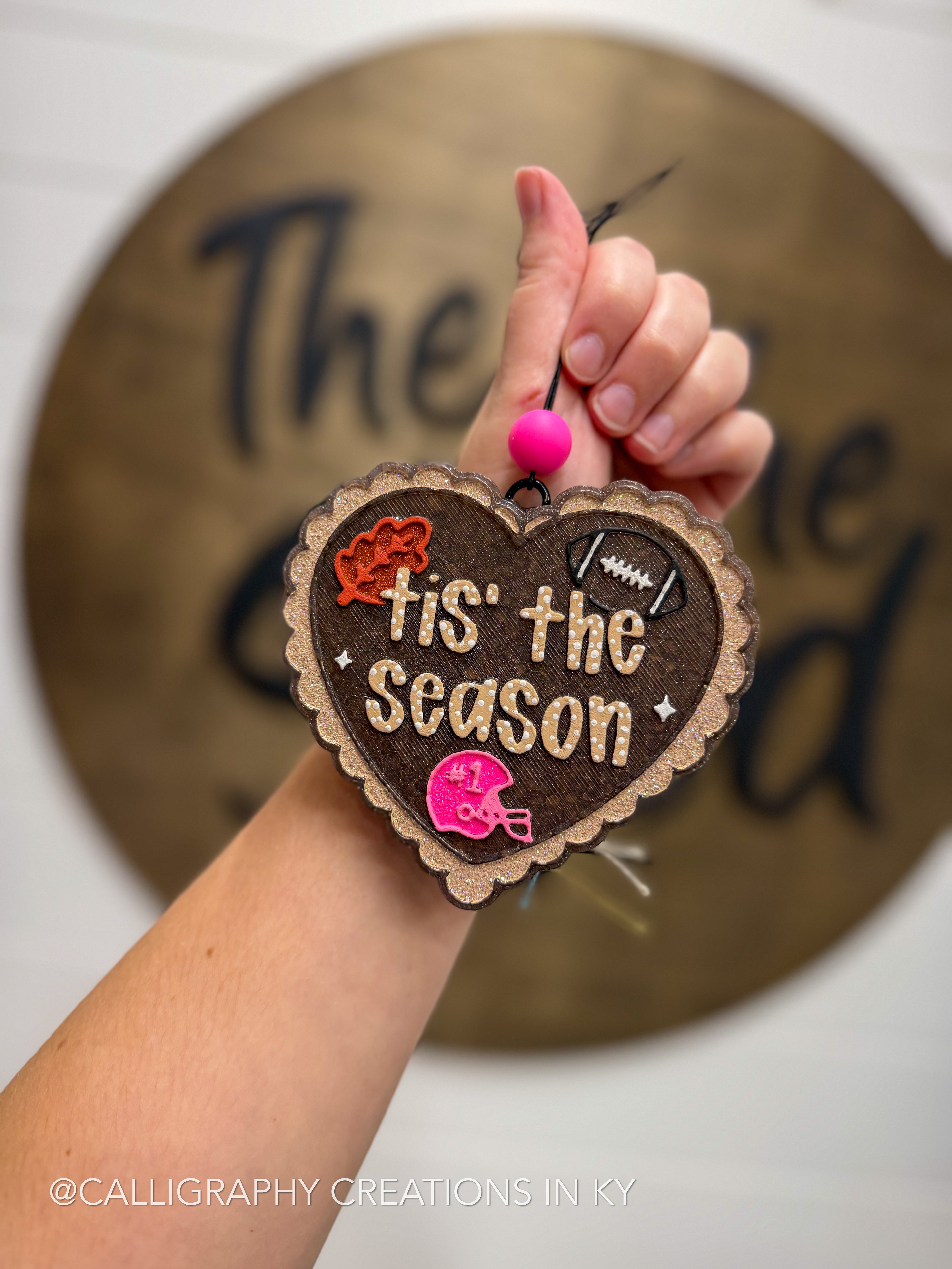 A hand holds a heart-shaped freshie with the phrase 'tis the season' in raised lettering. The ornament is decorated with small football-themed embellishments, including a football, helmet, and leaves, along with bright pink pom-poms.
