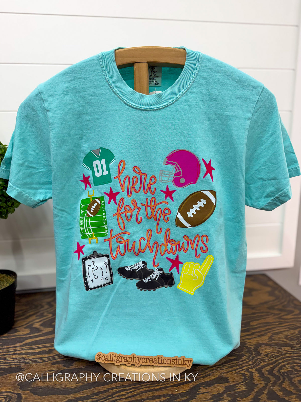 A turquoise short-sleeve t-shirt on a wooden hanger, featuring a colorful graphic design with football-themed illustrations and the text 'here is for the touchdowns'.