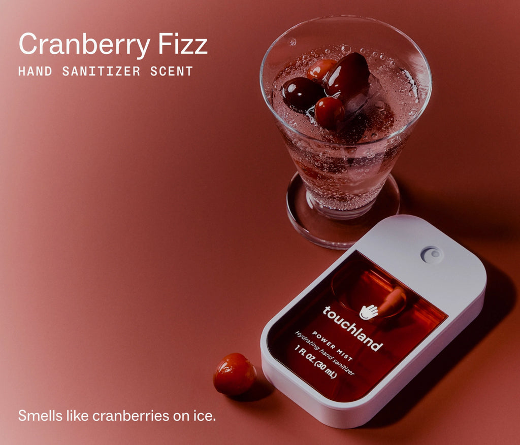 A hand sanitizer scent product shown on a warm reddish-brown background, featuring a white rectangular container labeled 'touchland' and 'Cranberry Fizz' alongside a glass of cranberry soda and cranberry pieces. Text on the image reads 'Cranberry Fizz HAND SANITIZER SCENT' and 'Smells like cranberries on ice.