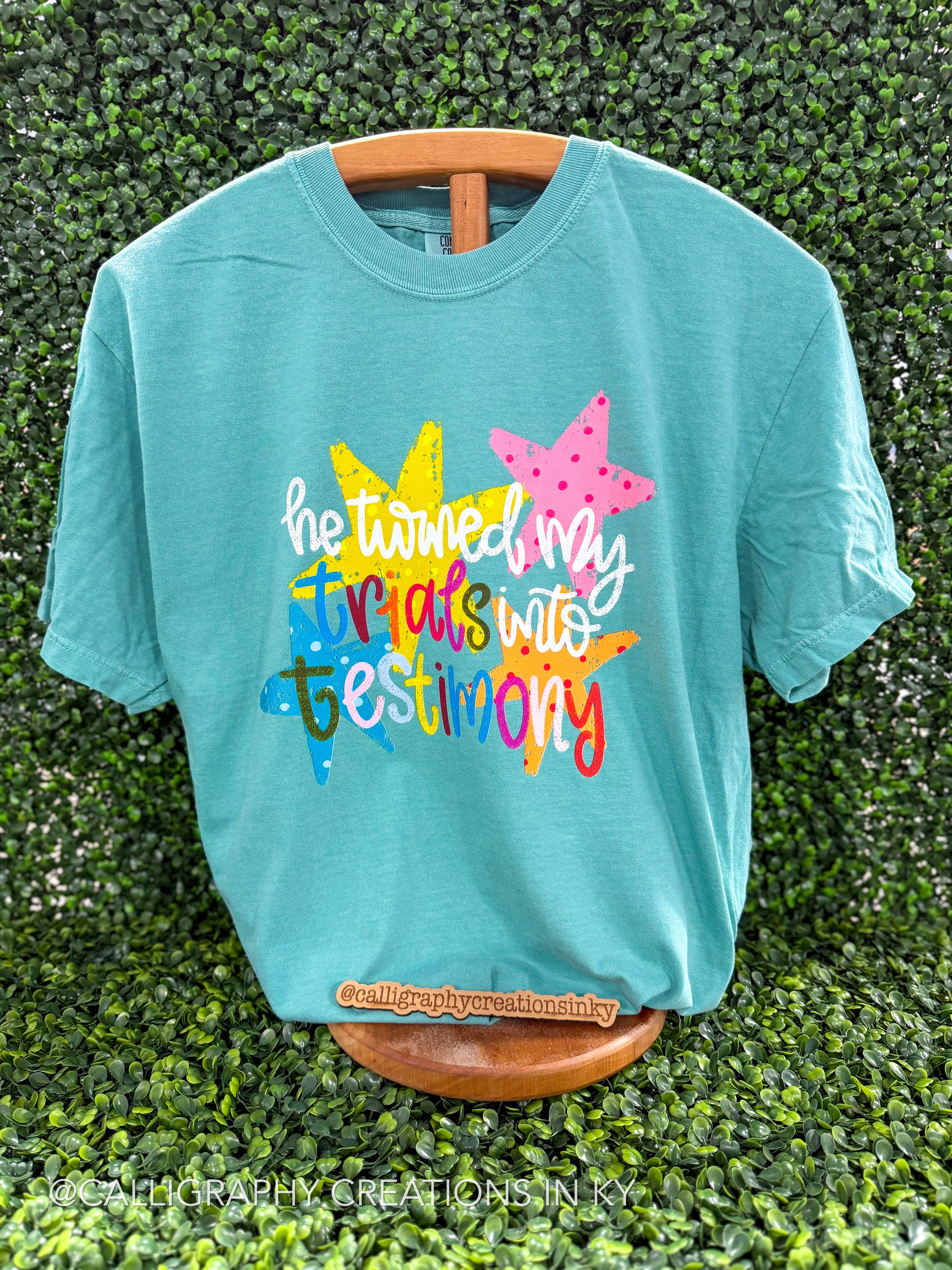 A turquoise short-sleeve graphic T-shirt displayed on a wooden hanger against a green leaf wall. The shirt features a colorful graphic with two star shapes and the text 'he turned my trials into testimony' in white and multicolored lettering.