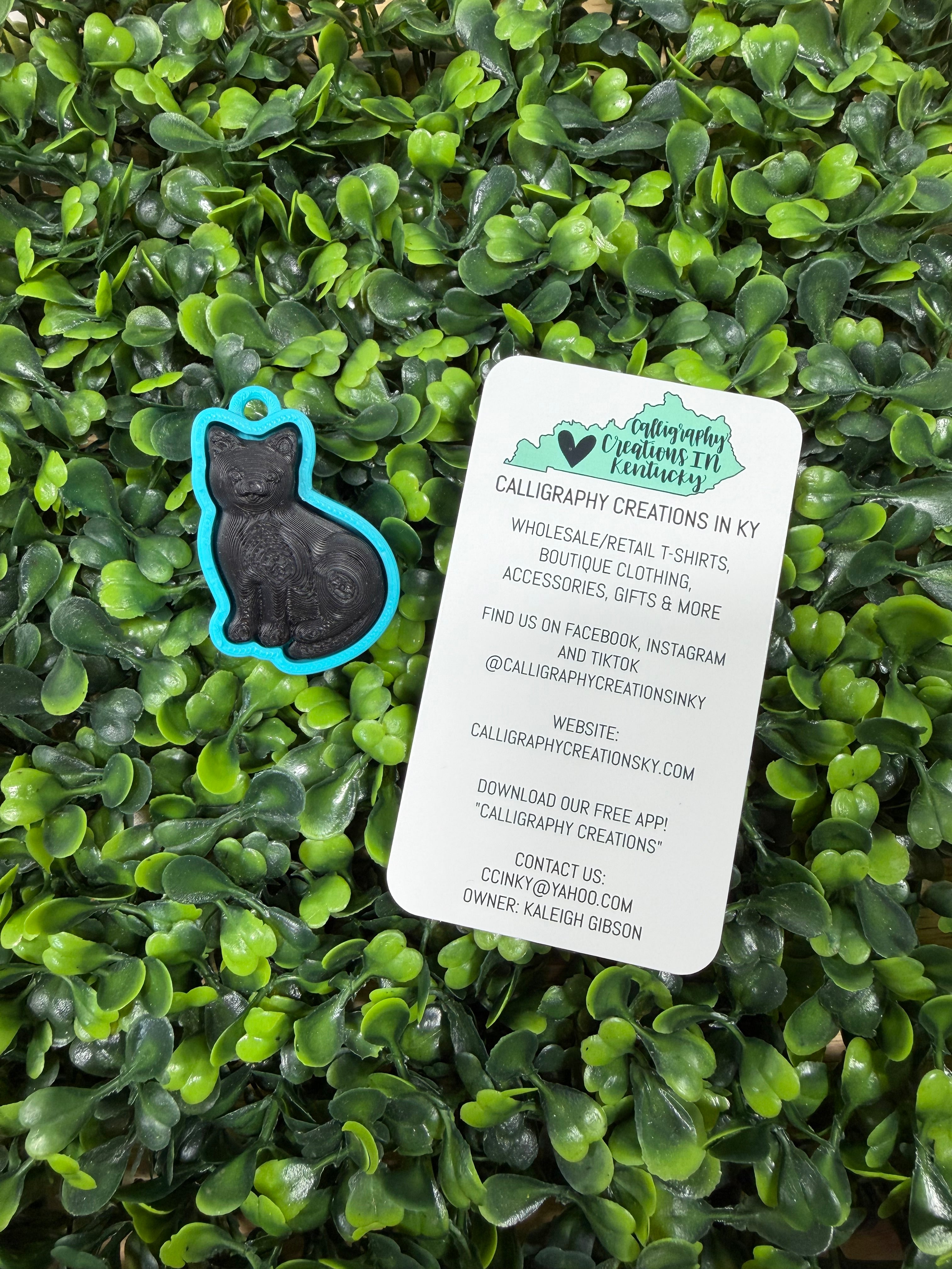 A black metal cat silhouette charm with a bright blue border and a small blue loop at the top, placed on a bed of green leaves next to a white product information card.
