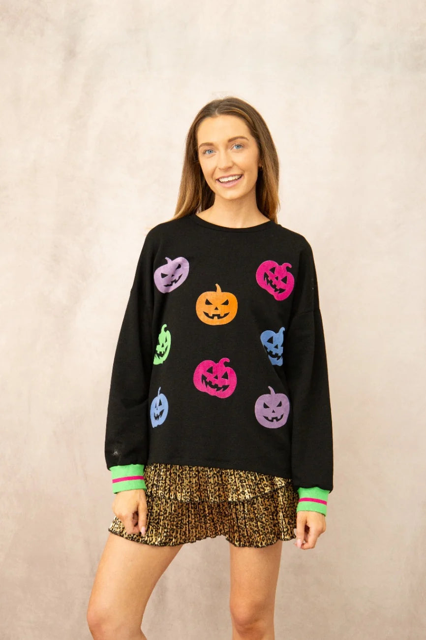 A woman modeling a black crewneck sweatshirt decorated with colorful embroidered jack-o'-lantern pumpkins in pink, orange, purple, green, and blue, paired with a leopard-print skirt against a textured beige background.