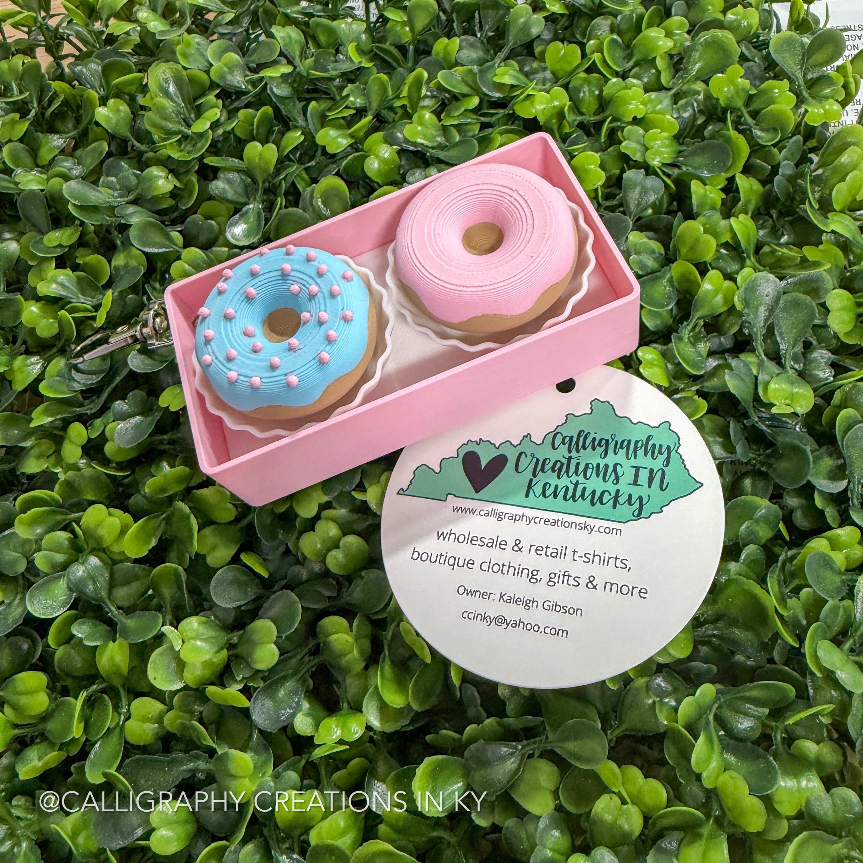Side view of a pink box with donut-shaped fidget clickers in the form of a keychain on a green leafy background, featuring Calligraphy Creations in Kentucky branding.