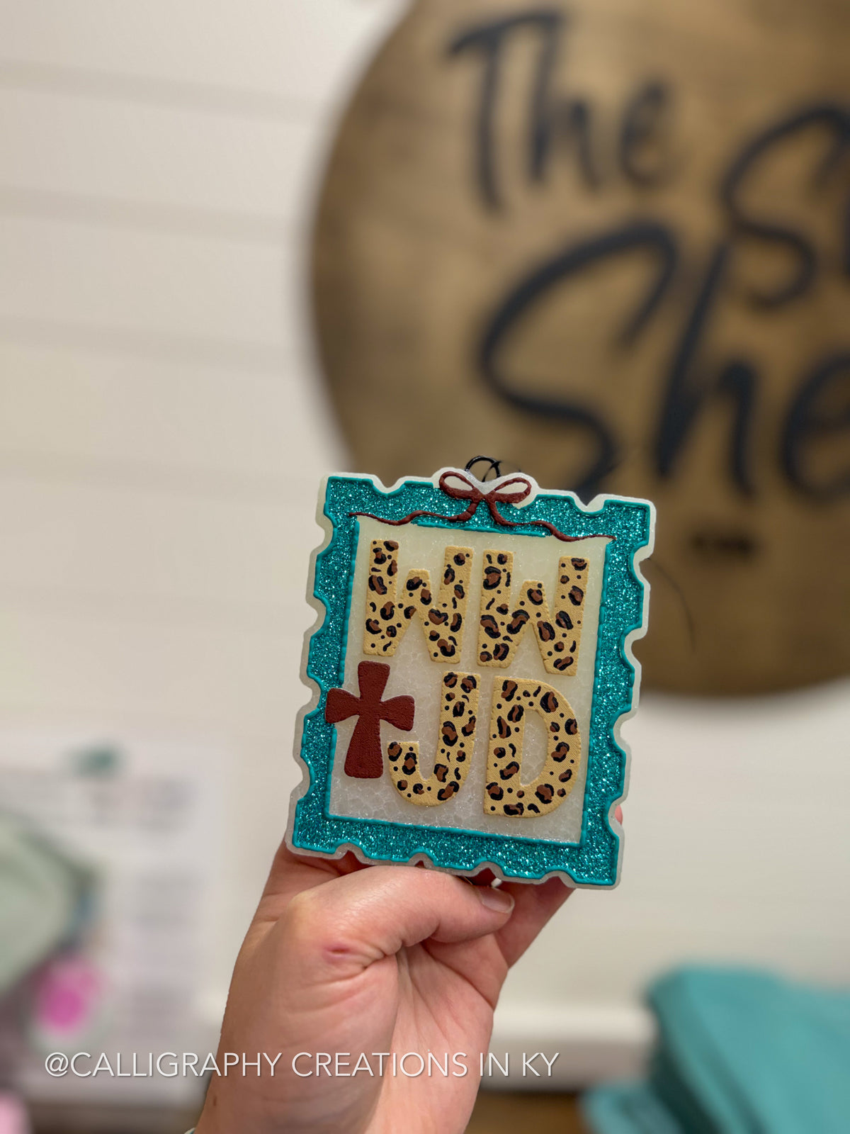 A hand holds a decorative freshie featuring the phrase 'WWJD’ in leopard-print letters inside a turquoise glitter frame with a red cross and bow detail. The background is a softly blurred indoor setting with a wooden sign.