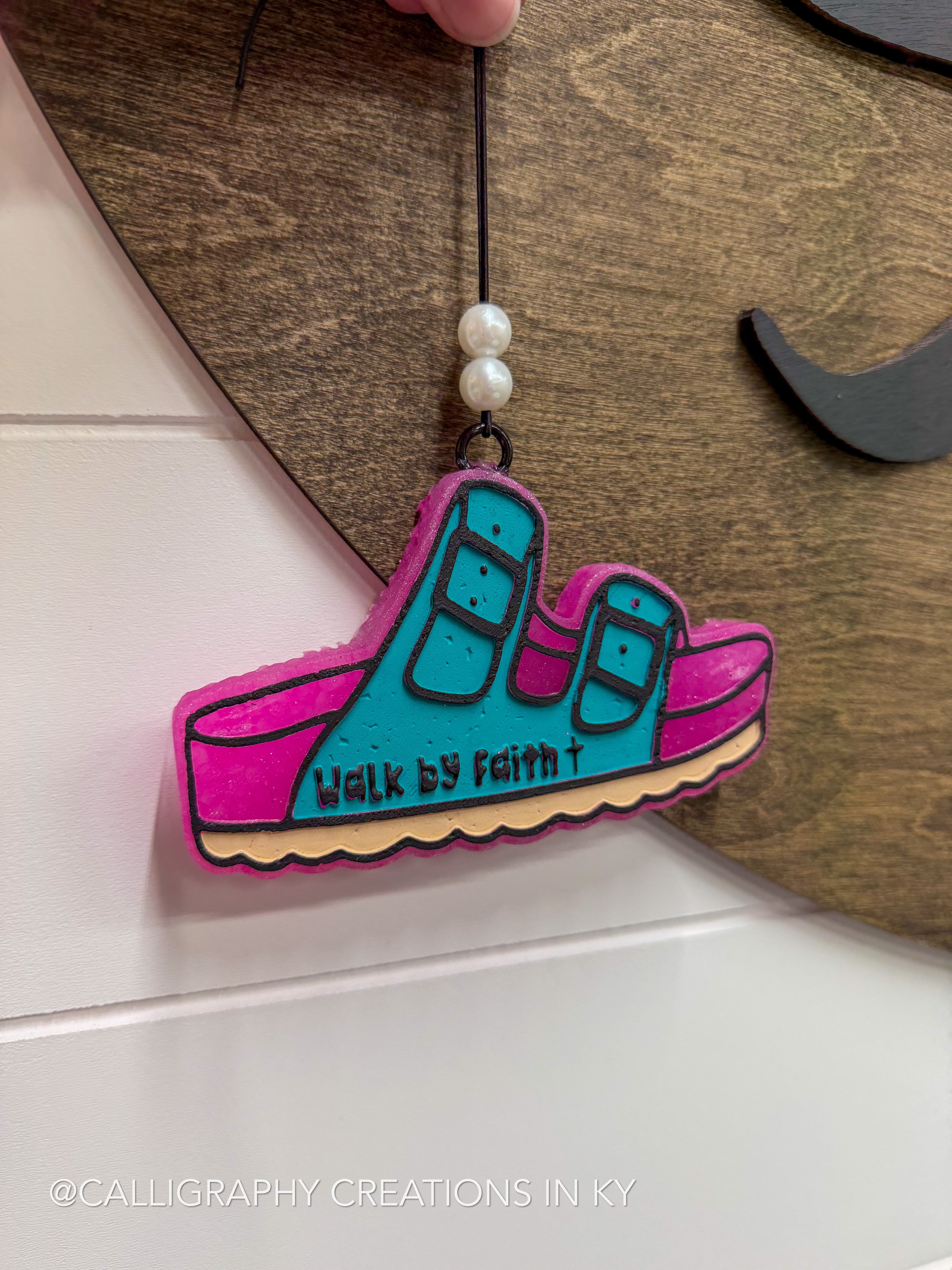 A colorful wooden shoe-shaped ornament with the words 'Walk by Faith' written on it, hanging from a black cord with white beads. The ornament features a turquoise upper, bright pink sole, and a beige wavy bottom edge, set against a dark wood background.