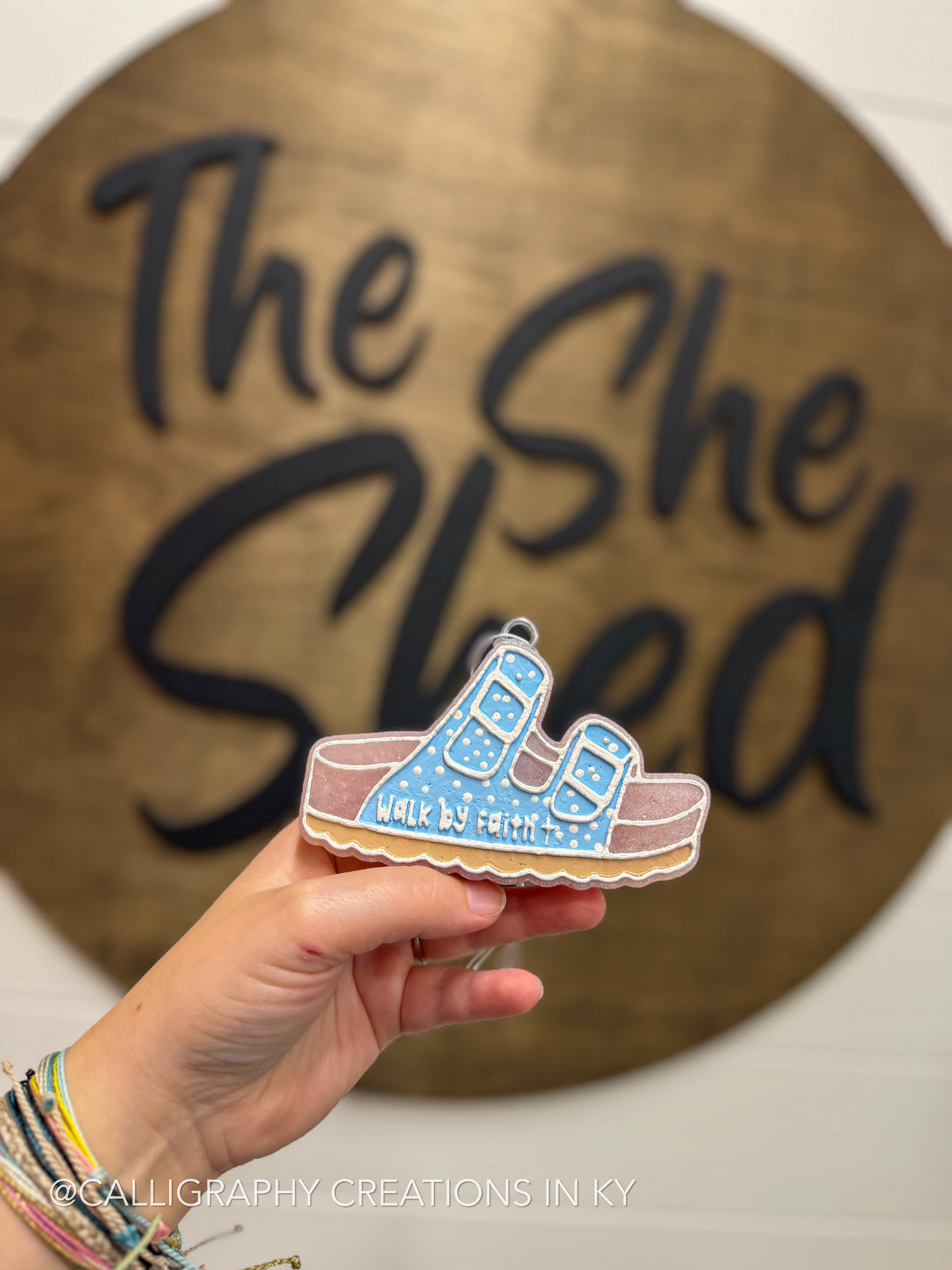 A hand holds a freshie shaped like a pair of blue sandals on a pink sole, with the words 'WALK BY FAITH' on the front. The background shows a wooden sign that reads 'The She Shed' in black script.