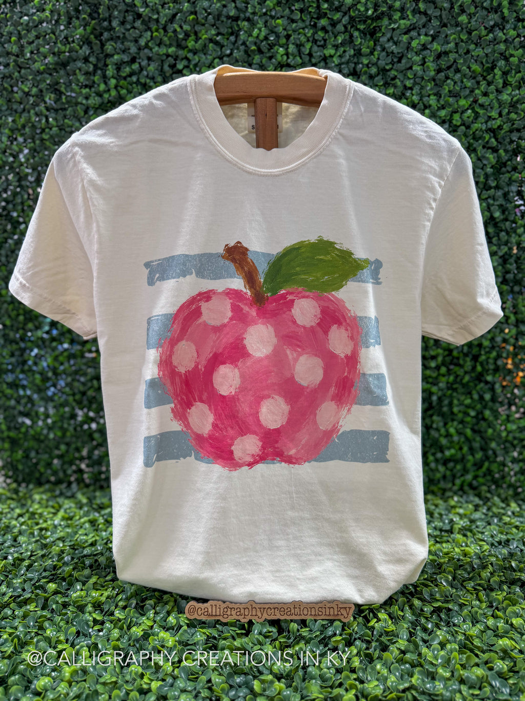 A white short-sleeve t-shirt displayed on a wooden hanger against a green leafy hedge background. The shirt features a painted graphic of a pink apple with white polka dots, a green leaf, and light blue horizontal stripes.