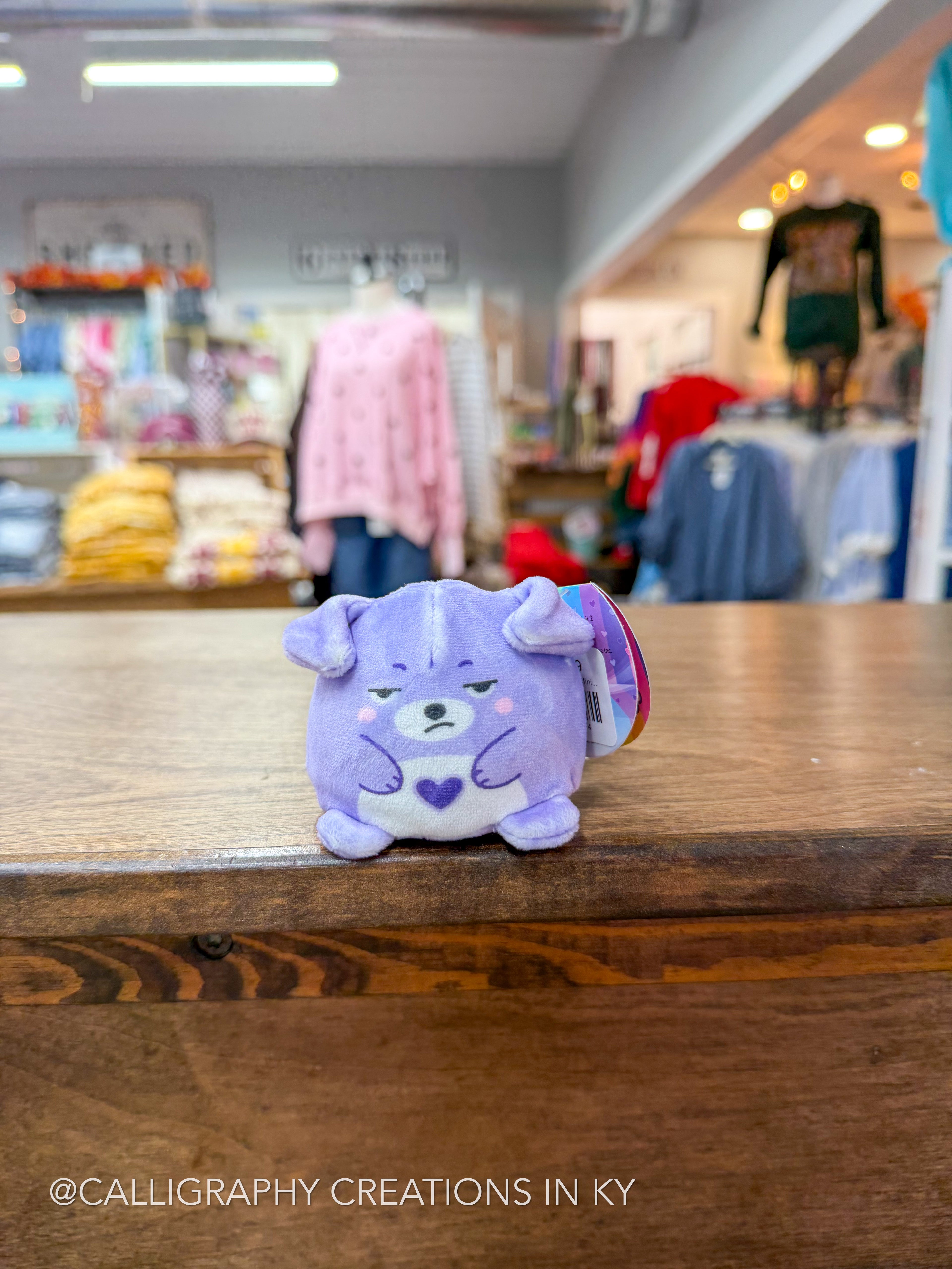 A small purple plush toy with a white snout and chest, featuring a heart on its chest, sitting on a wooden counter in a blurred retail store setting.
