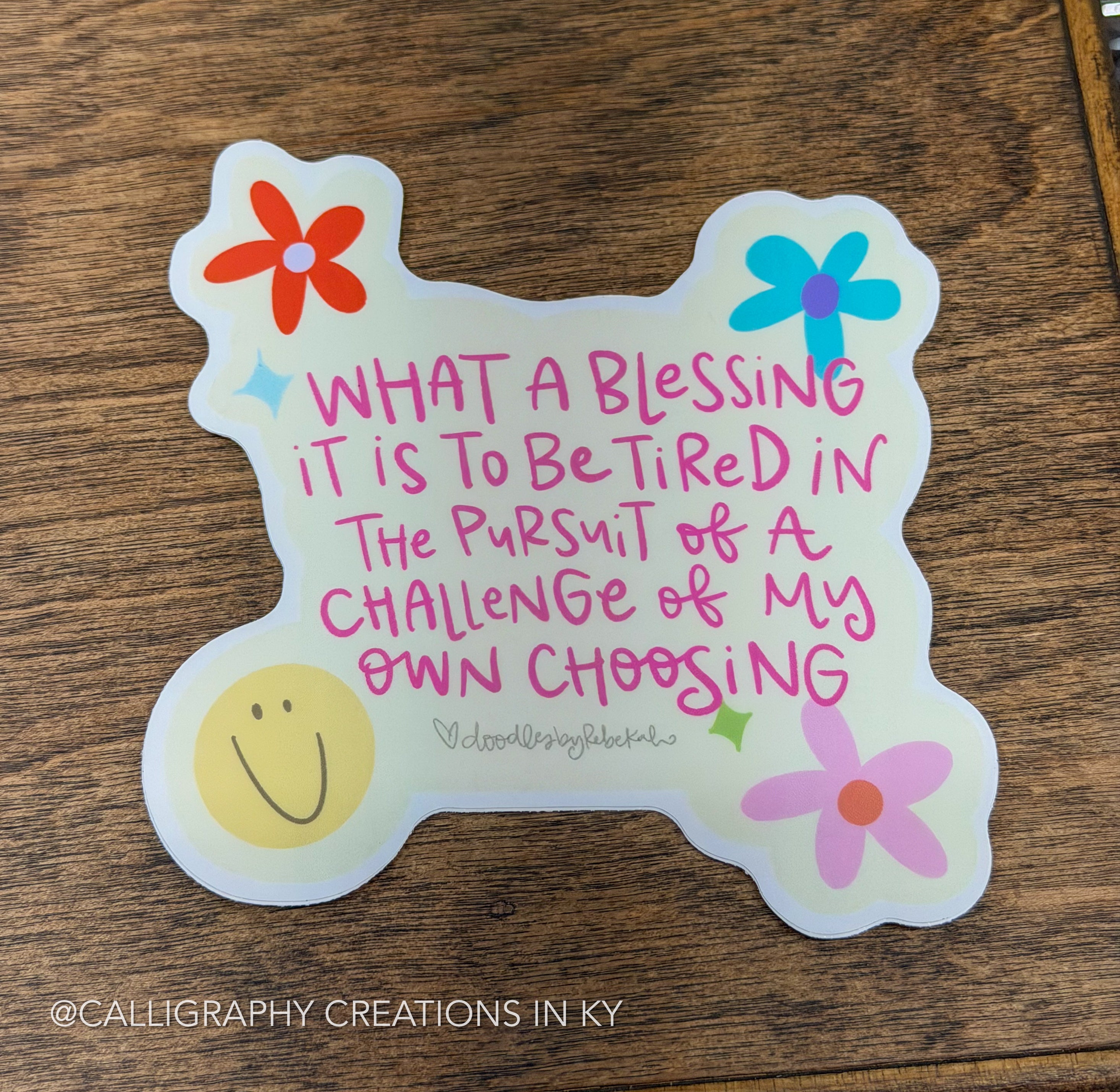 A die-cut sticker with a whimsical bone shape, featuring a handwritten quote in pink and black ink, decorated with colorful flowers and a smiley face, displayed on a wooden surface.
