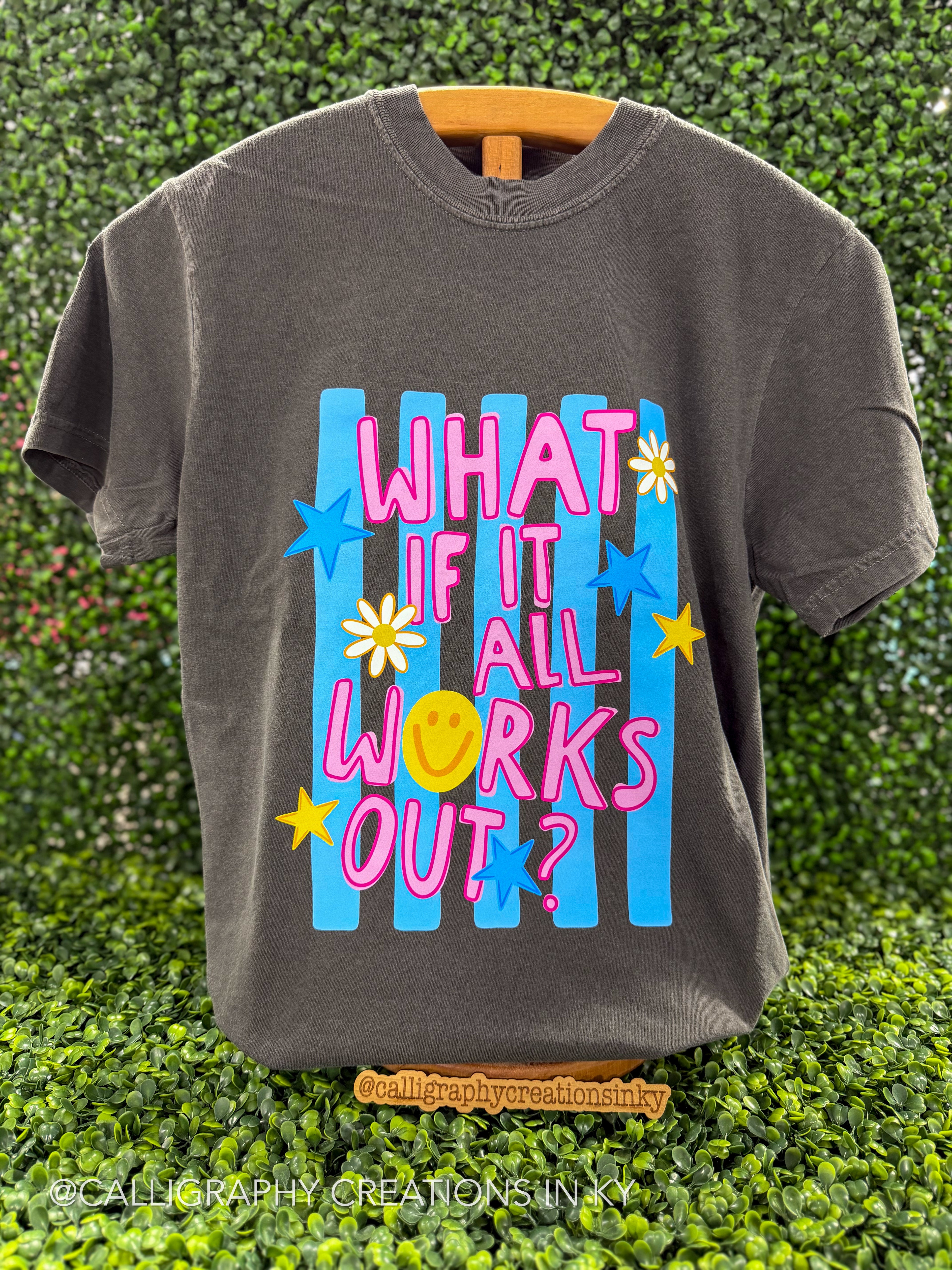 A charcoal gray graphic t-shirt hanging on a wooden hanger against a green hedge background. The shirt features a colorful retro-style graphic with the text 'WHAT IF IT ALL WORKS OUT?' in pink and yellow, set against blue vertical stripes and decorated with blue and yellow stars and white daisy flowers.