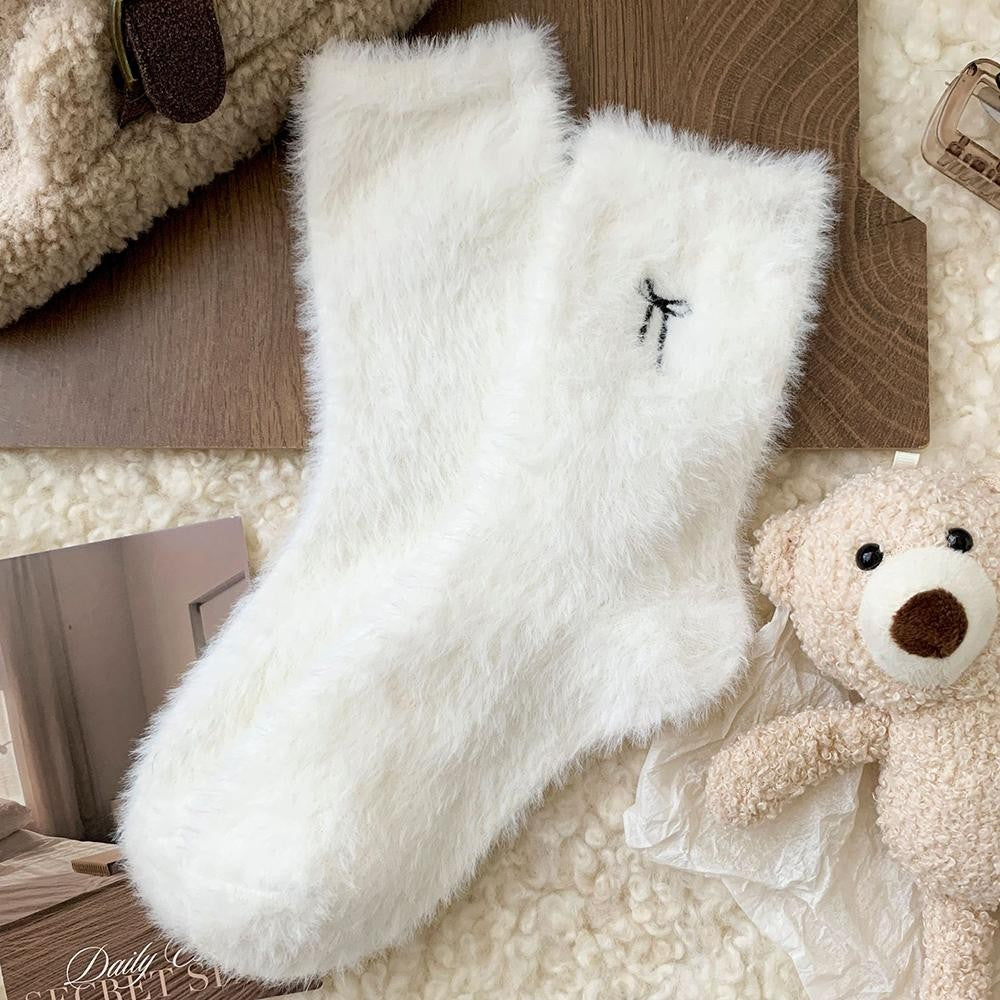 A pair of fluffy white ankle socks laid flat on a cream sherpa surface with styled props, including a teddy bear, wooden boards, and a textured bag. small black embroidered bow near the cuff.