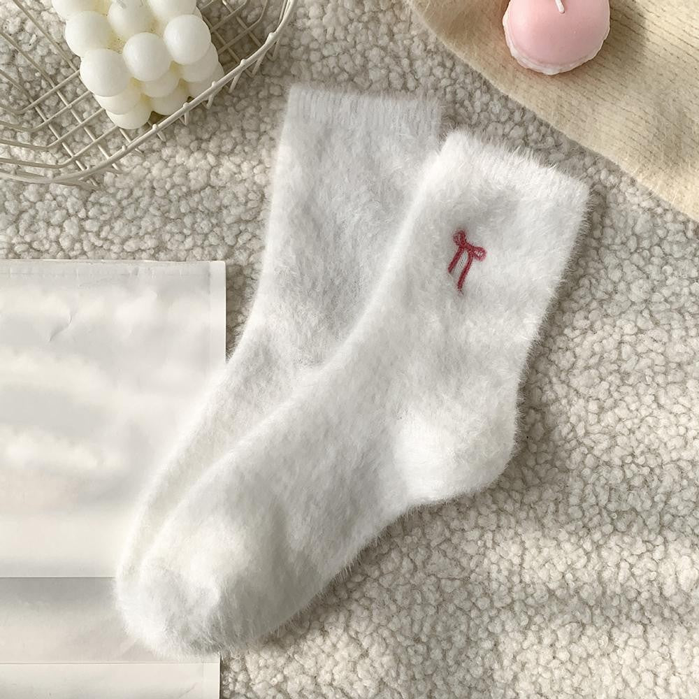 A pair of fluffy white socks with a small pink bow embroidery on the cuff, displayed on a textured beige surface with decorative props.