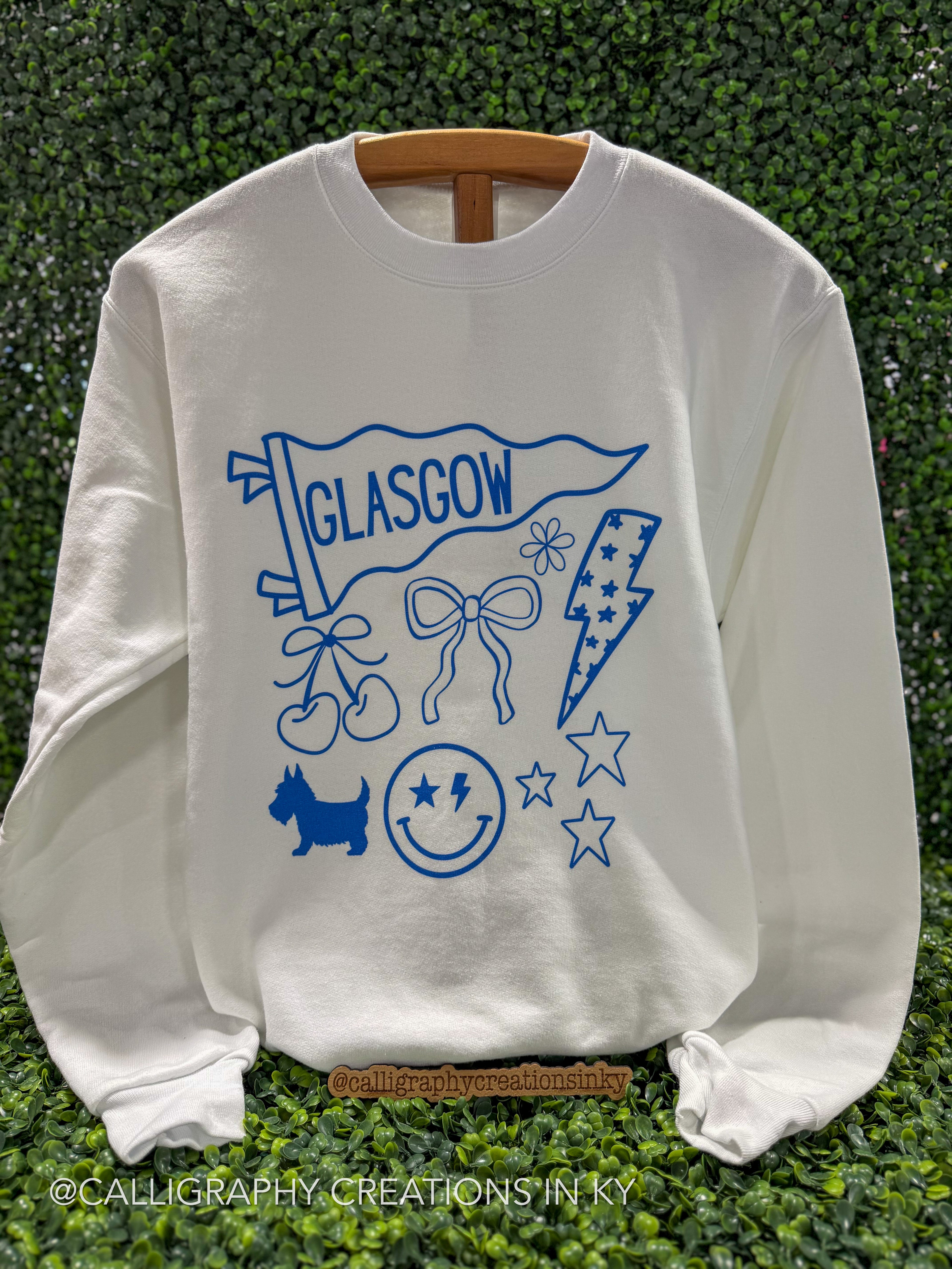 A white crewneck sweatshirt with a blue graphic print featuring the word 'GLASGOW', a flag, cherries, bows, a lightning bolt, a dog silhouette, stars, and a smiley face, displayed on a wooden hanger against a green hedge background.
