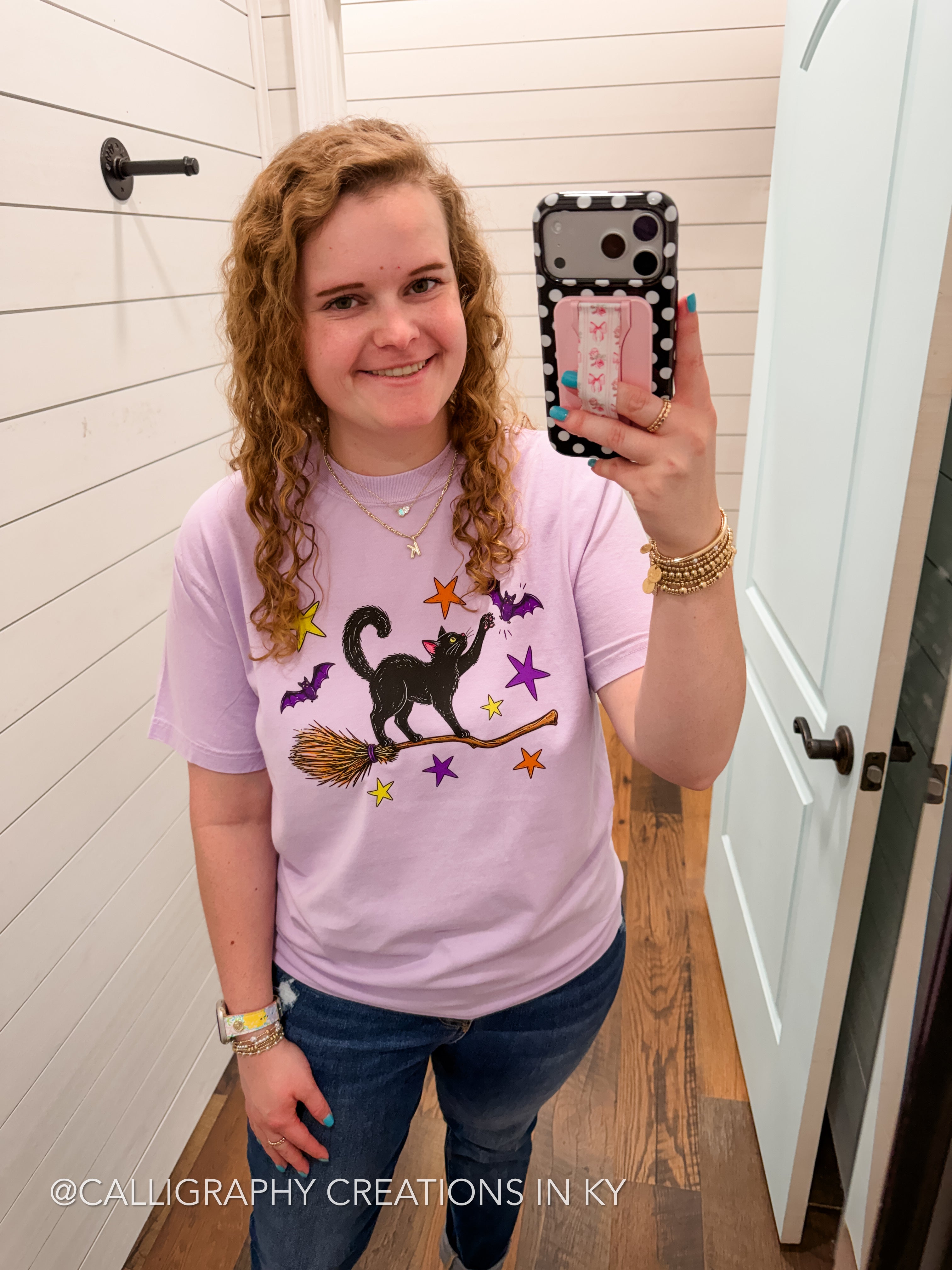 A woman takes a mirror selfie wearing a light purple t-shirt with a black witch cat graphic on a broomstick surrounded by purple and orange stars, paired with blue jeans.