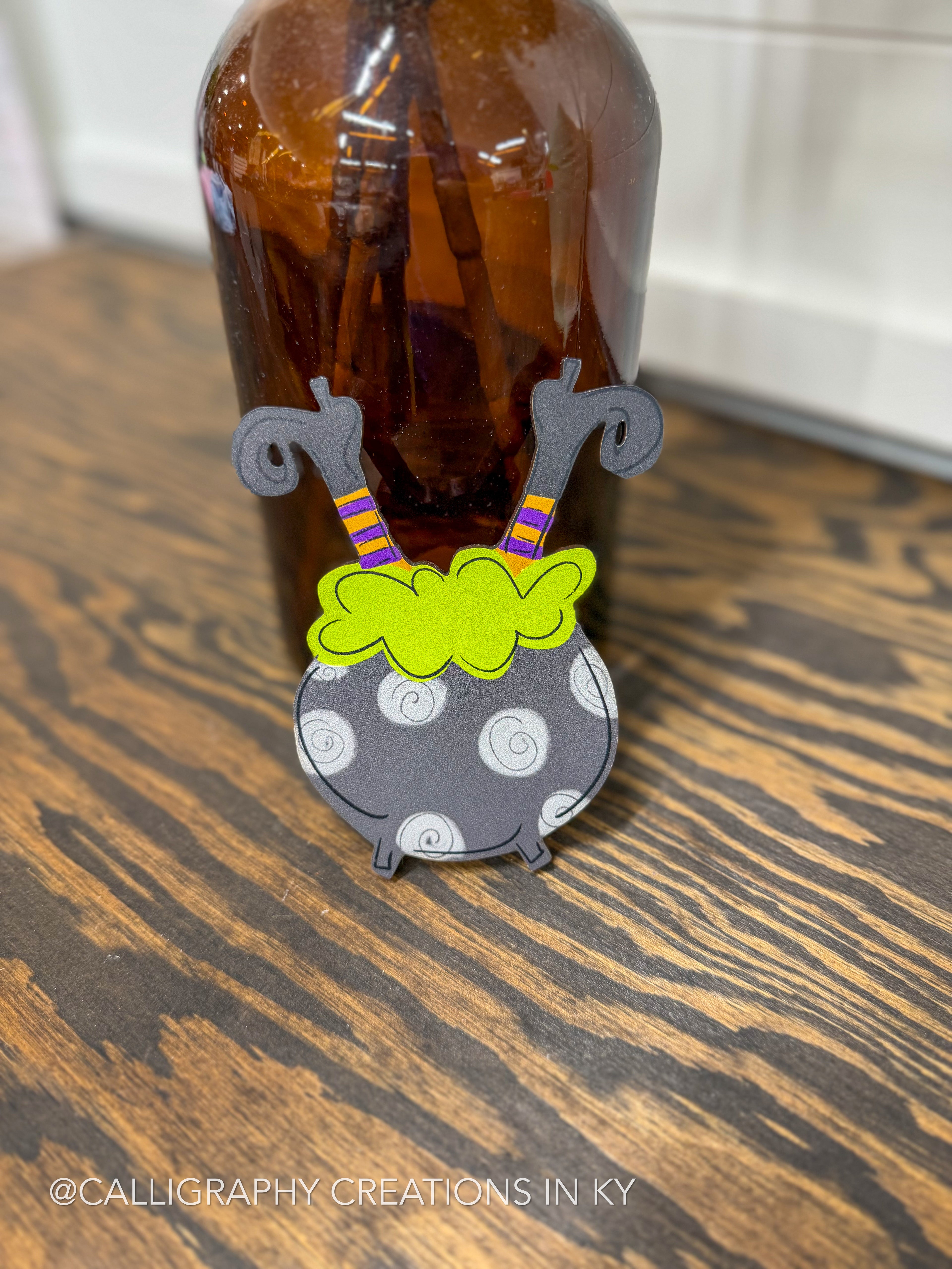 A small decorative calligraphy piece shaped like a witch's cauldron with gray legs, purple and yellow striped socks, a bright green top, and a gray body with white swirls, displayed on a wooden surface in front of an amber glass bottle.