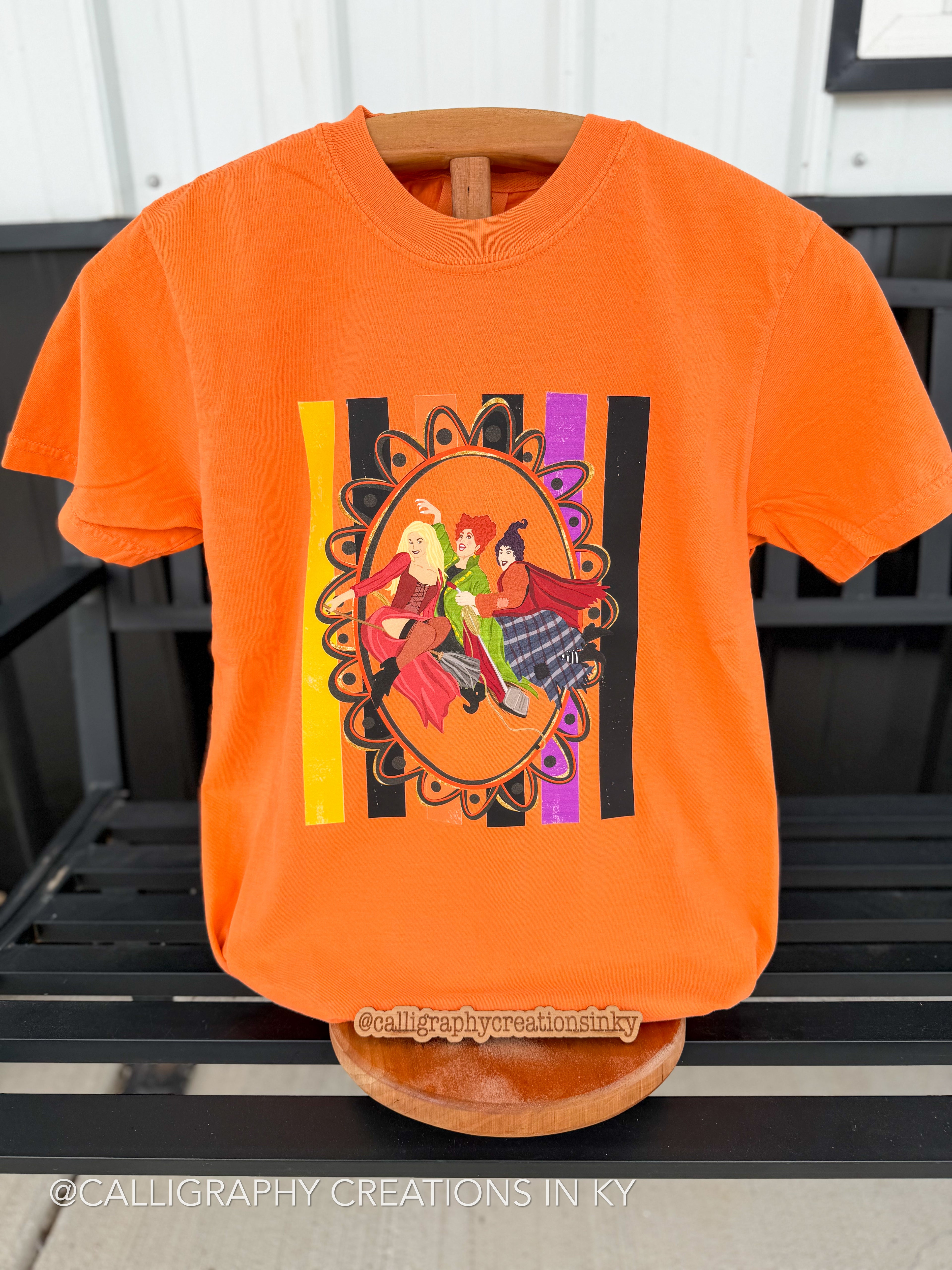 An orange short-sleeve graphic T-shirt displayed on a wooden hanger, featuring a colorful central print with a woman in a red dress and a woman in a green dress, set against black and yellow vertical stripes.