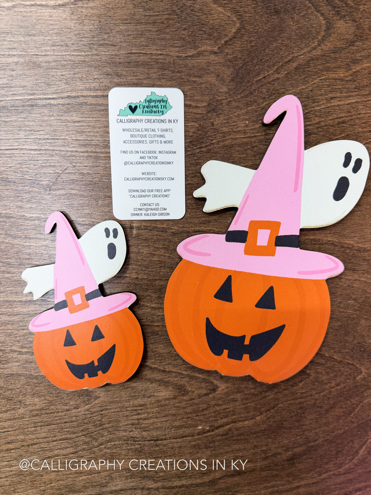 Two orange Halloween pumpkin cutouts wearing pink witch hats with black trim, displayed on a brown wood background with a white Calligraphy Creations business card.