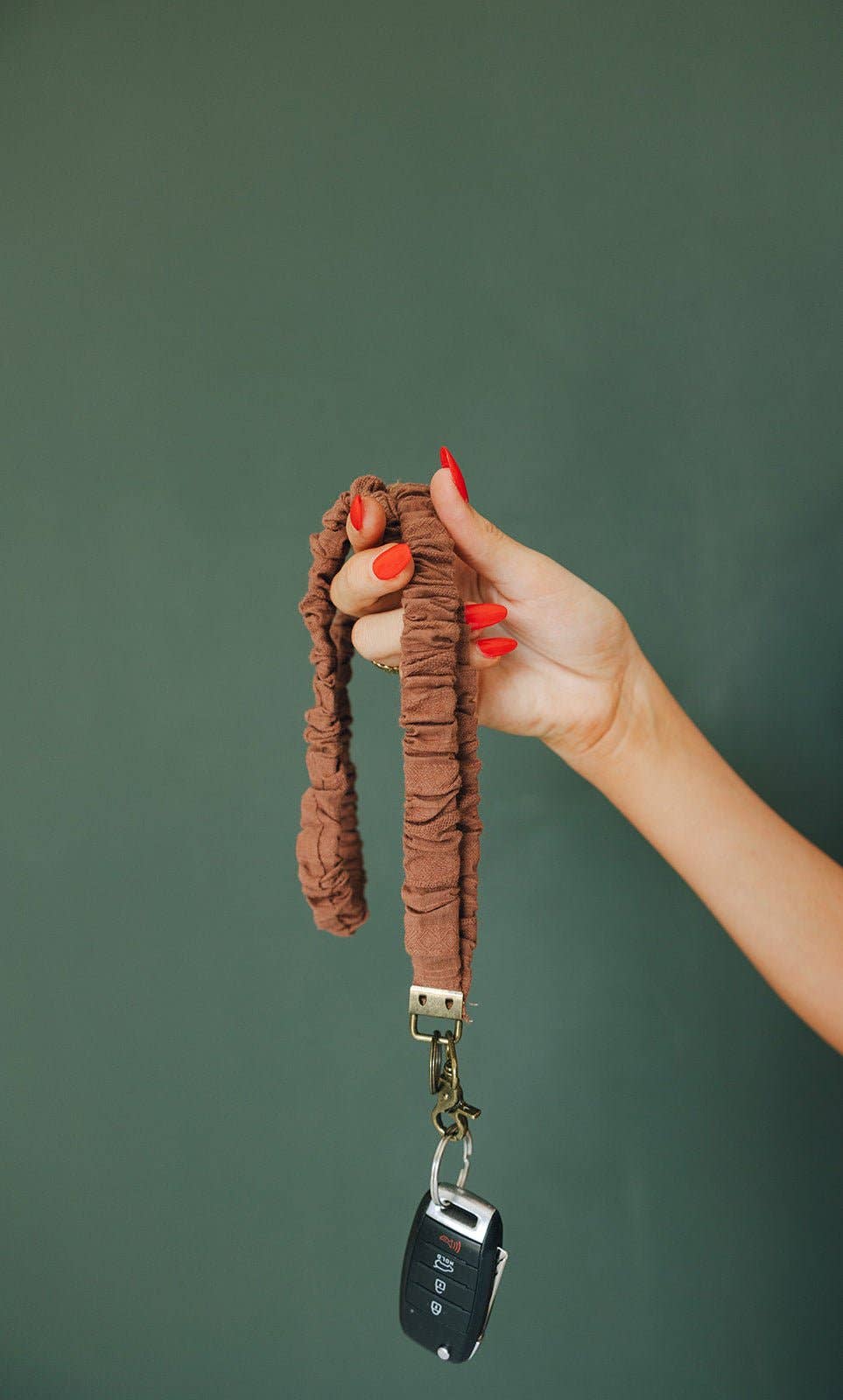 Person holding brown scrunchie keychain with gold clasp and key fob attached against a green background 