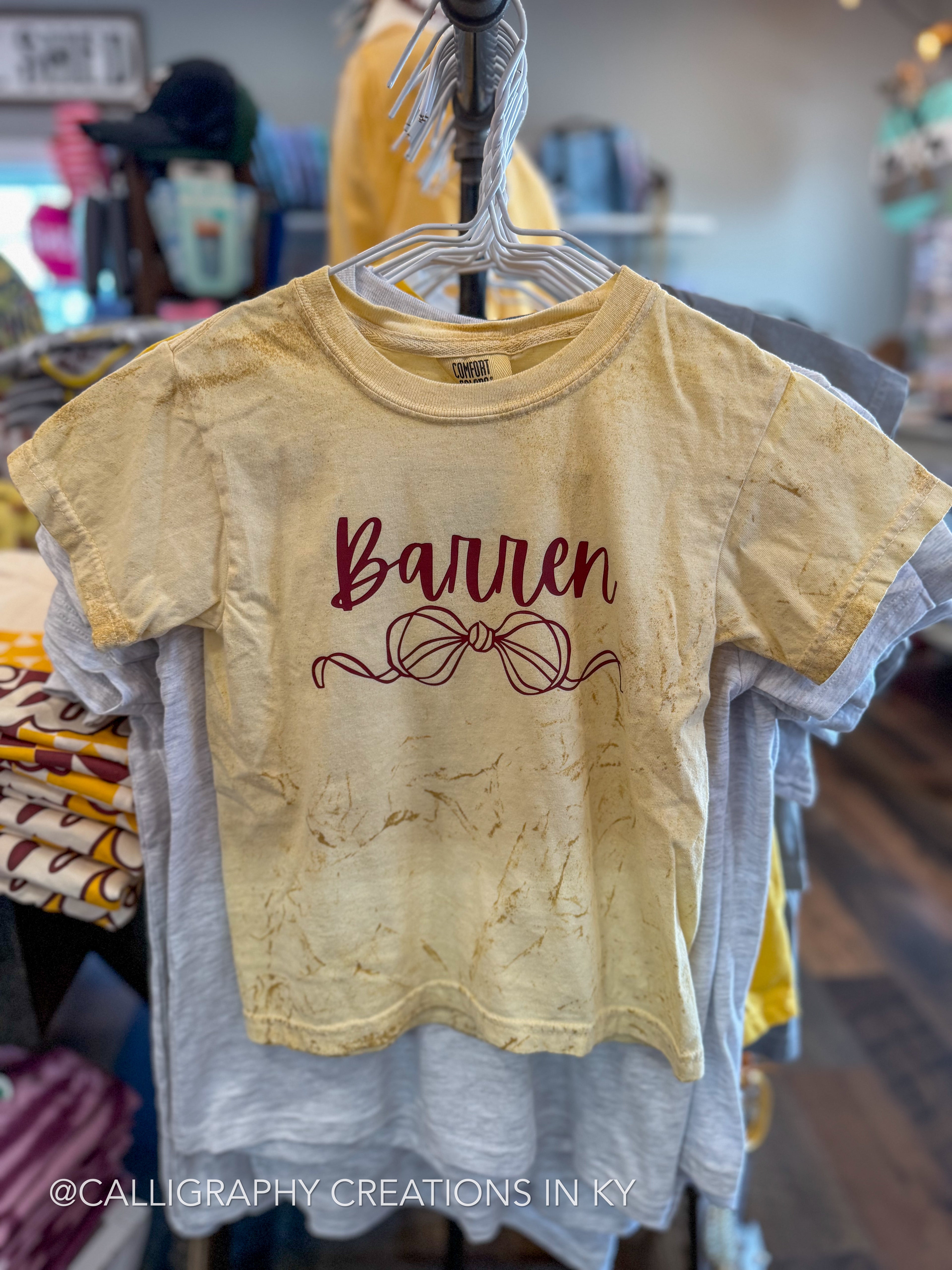 A short-sleeve graphic t-shirt in a warm, faded yellow-beige color, hanging on a hanger in a retail store. The shirt features the word 'Barren' in dark burgundy cursive script with a matching ribbon/bow illustration underneath.