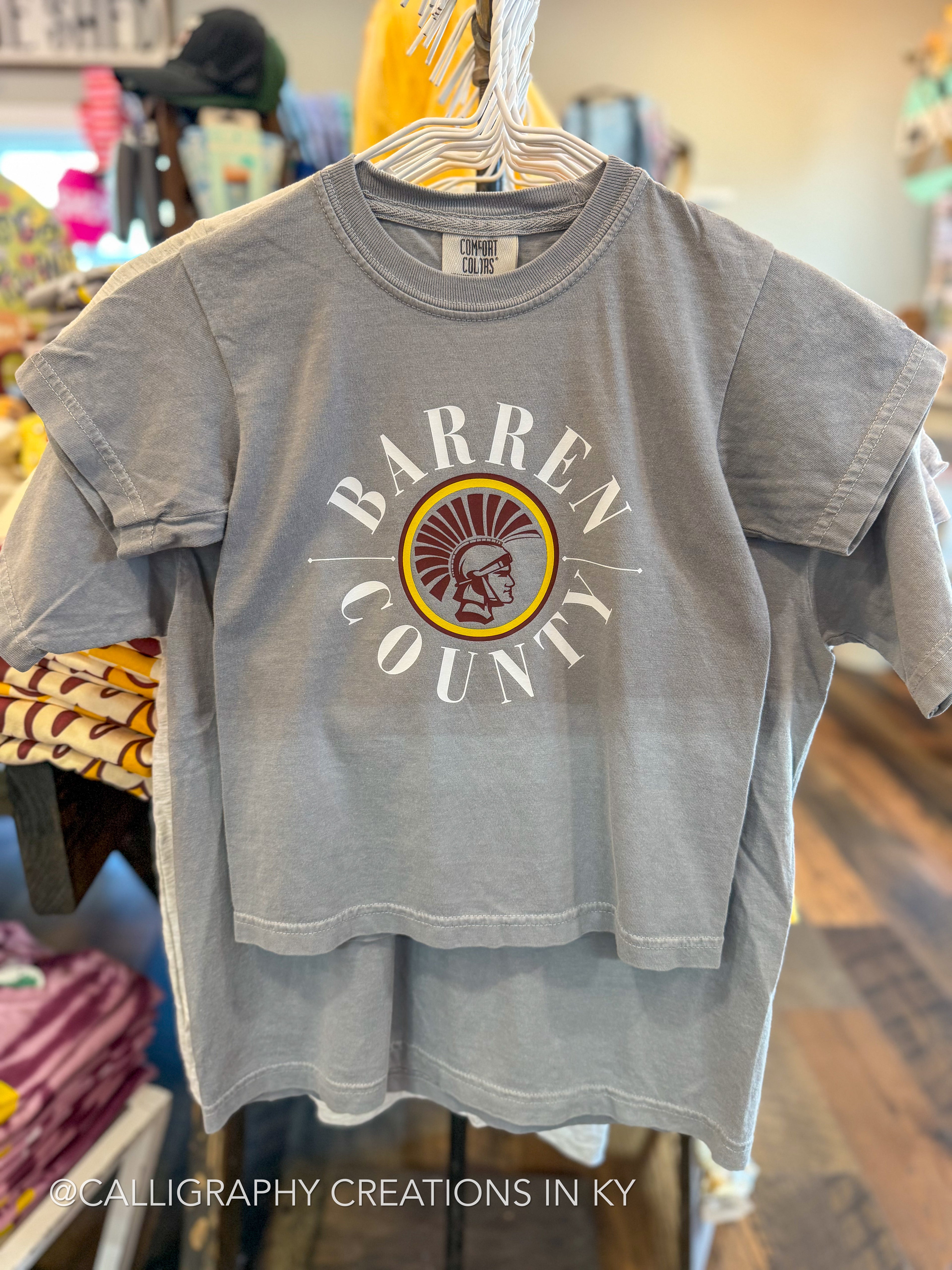 A gray short-sleeve t-shirt hanging on a hanger in a retail store, featuring a distressed graphic on the front that reads 'BARREN COUNTY' in white around a circular emblem with a Trojan head in yellow, red, and brown.
