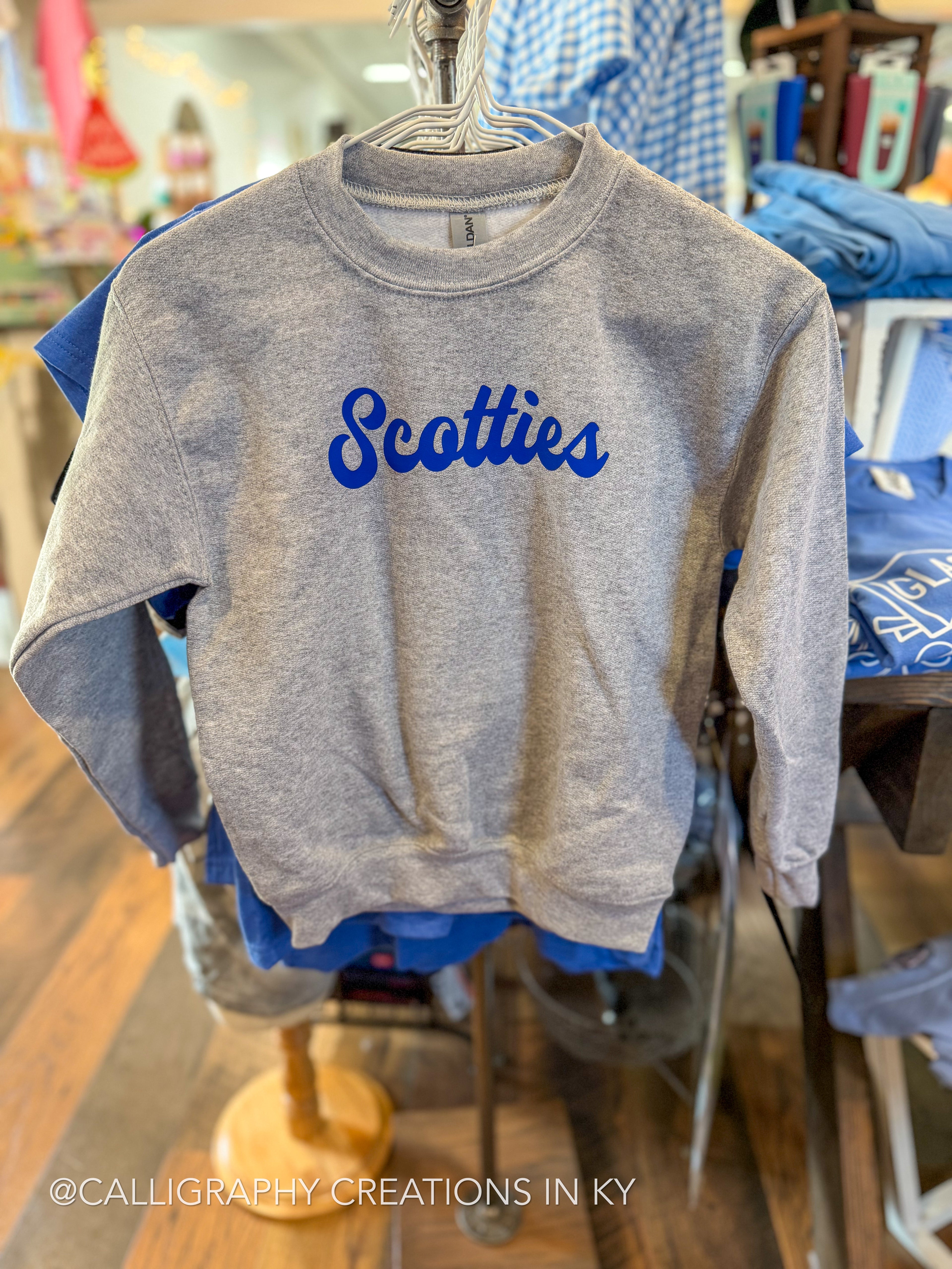 A heather gray crewneck sweatshirt hanging on a metal hanger, featuring the word 'Scotties' printed across the chest in blue cursive script. The sweatshirt is displayed in a retail store setting with other blue apparel visible in the background.