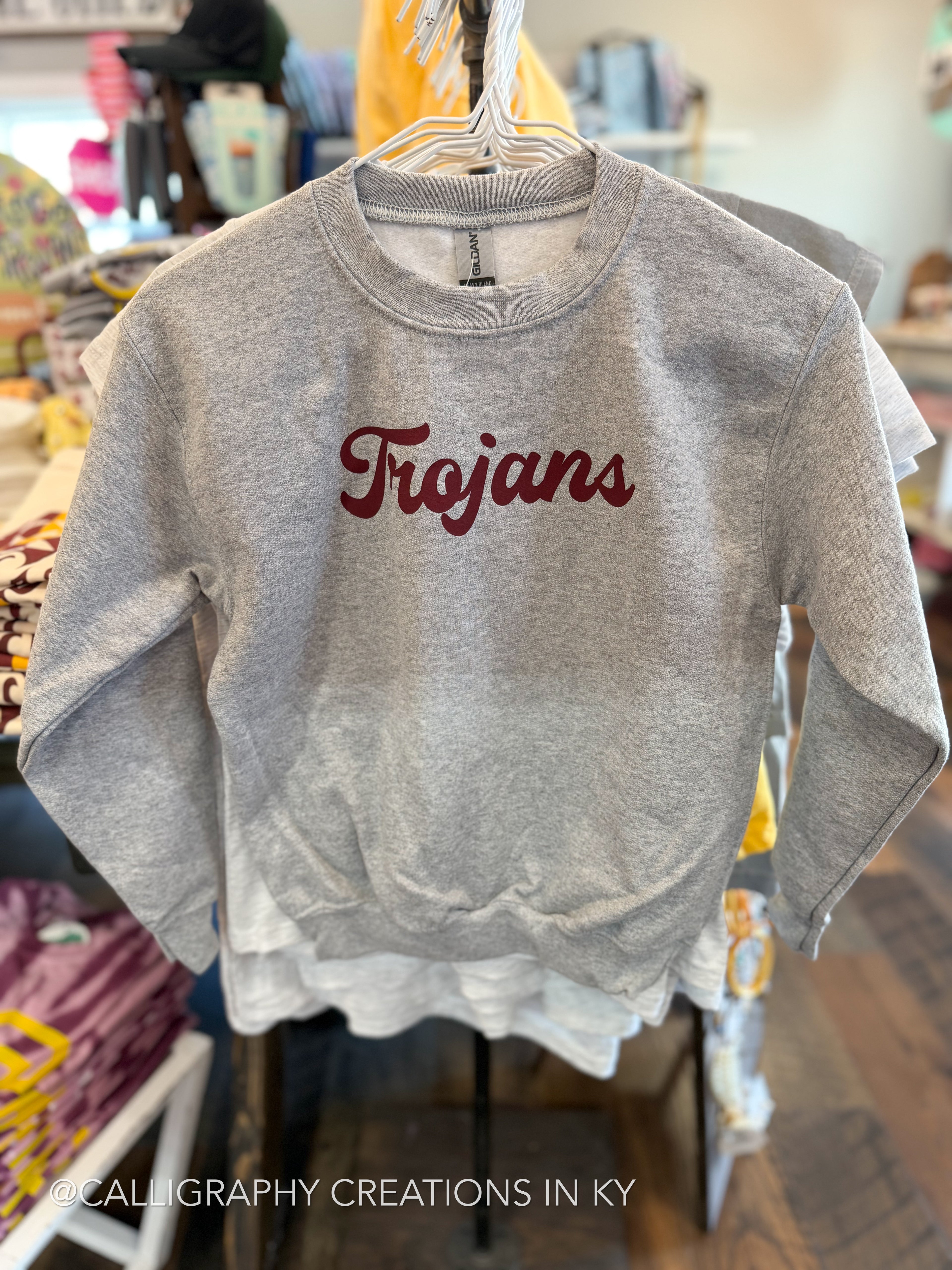 A heather gray crewneck sweatshirt with 'Trojans' written across the chest in dark red script, hanging on a white hanger inside a retail store.