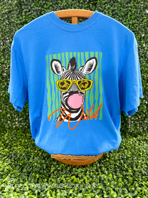 A bright blue graphic t-shirt displayed on a wooden hanger against a green hedge background. The shirt features a cartoon zebra wearing yellow sunglasses and blowing a pink bubblegum bubble, with green vertical stripes behind it and orange script text below.