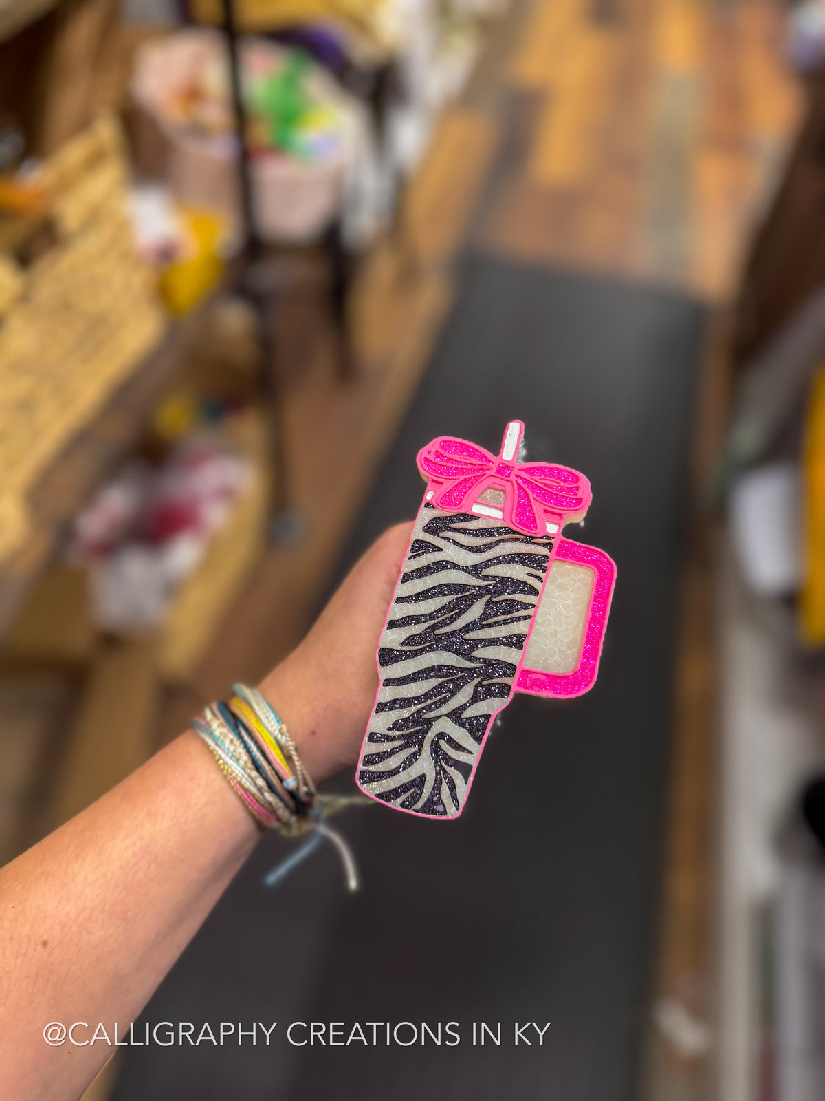 A hand holding a glittery pink and black zebra-patterned tumbler-shaped freshie with a pink bow, against a blurred indoor background.