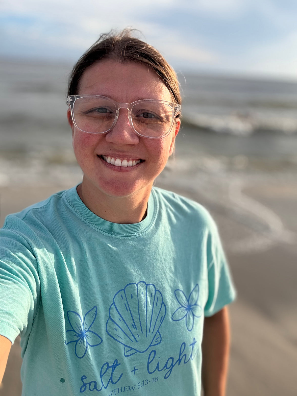 A smiling woman wearing clear eyeglasses and a chalky mint graphic t-shirt with blue seashell and floral designs, taking a selfie on a beach with the ocean waves blurred in the background.